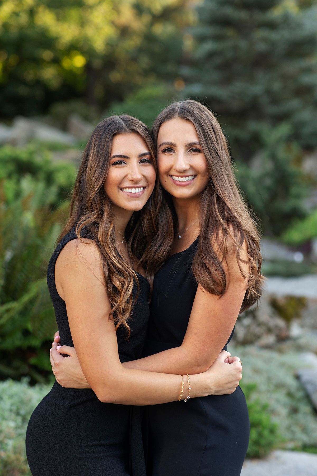 Two brunette women in black outfits smiling and hugging outdoors in a garden setting.