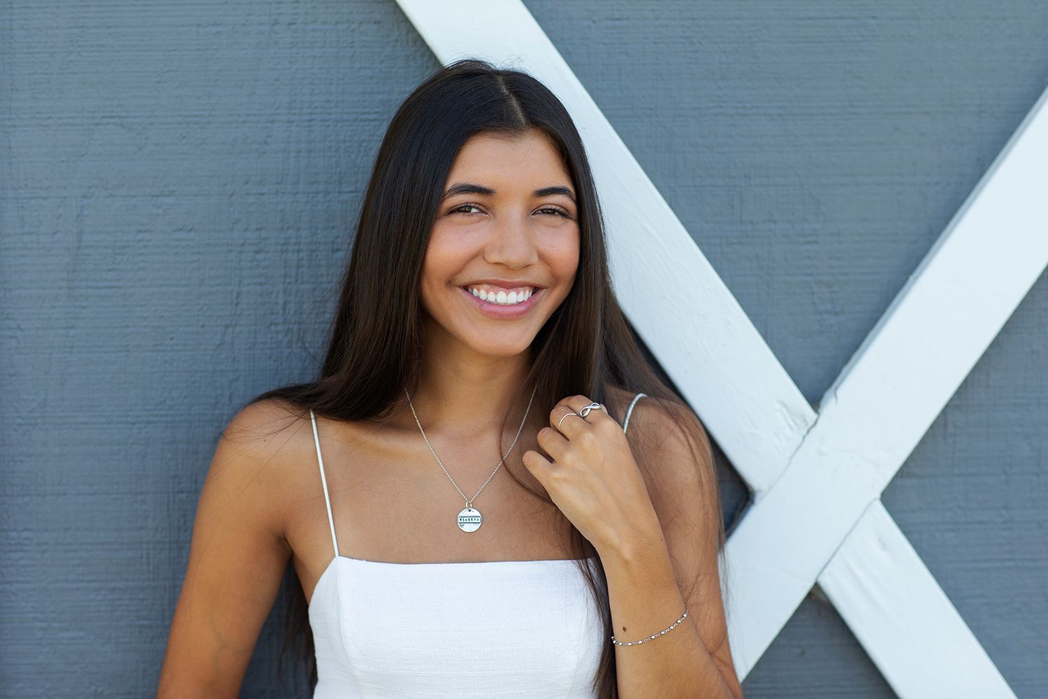Smiling young woman in white camisole wearing a delicate necklace, posing against a gray wall.
