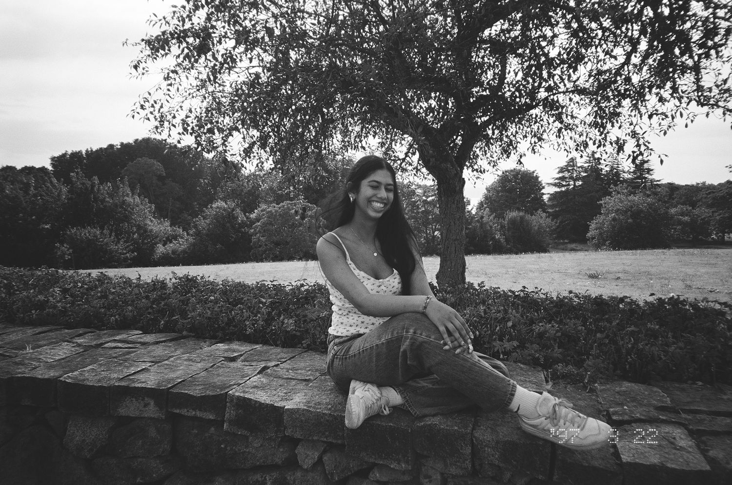 Smiling young woman sitting on stone wall in black and white photo with tree and garden behind her.