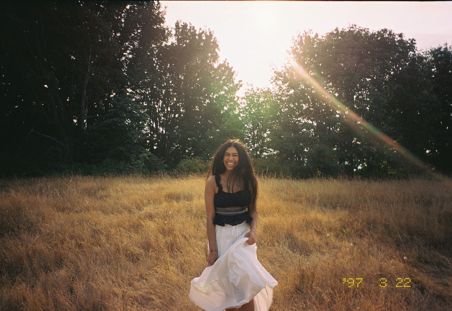 Film photo of young woman in black top and white skirt standing in golden field at sunset.