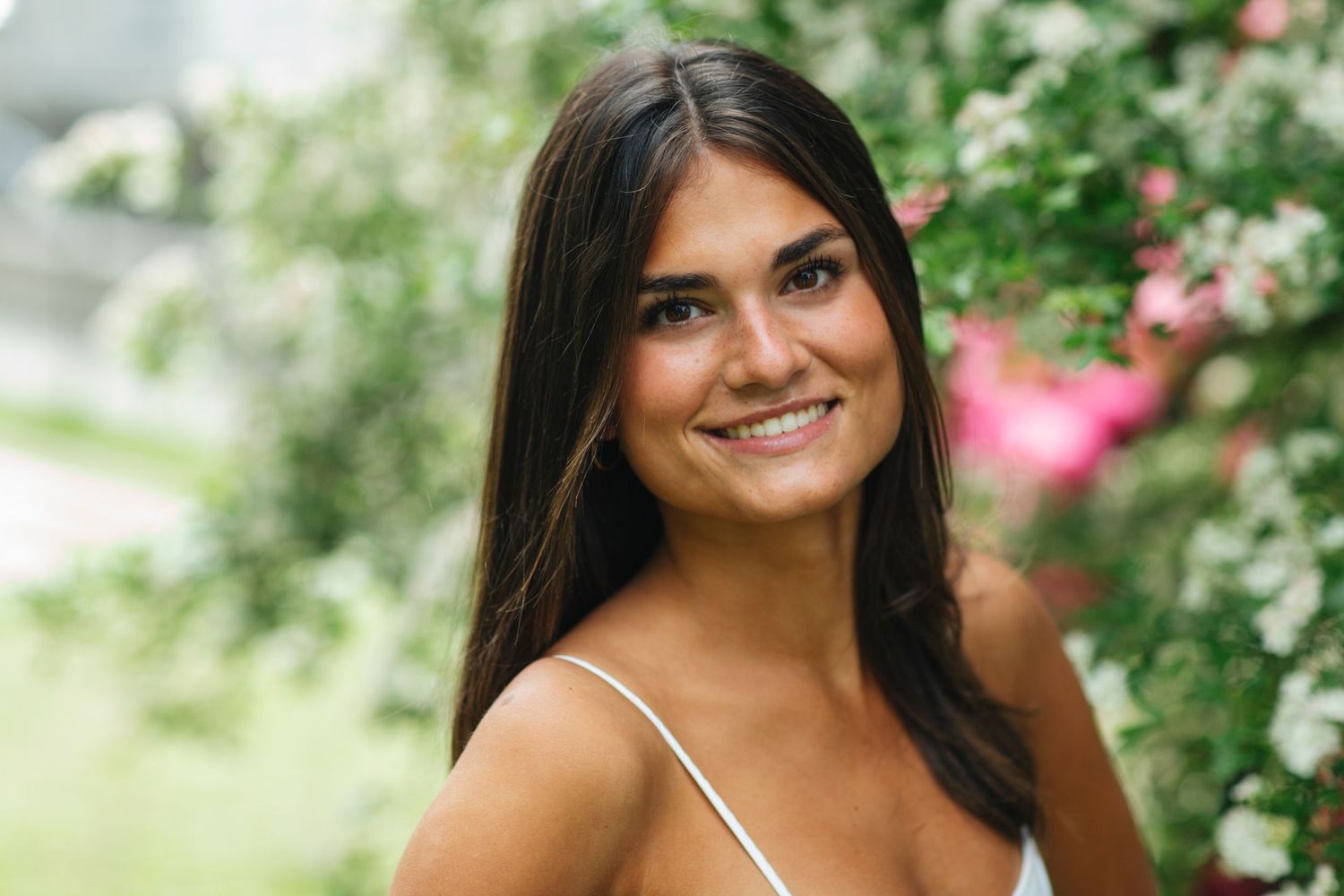 Smiling young woman with long dark hair in white top, posing outdoors near blooming pink and white flowers.