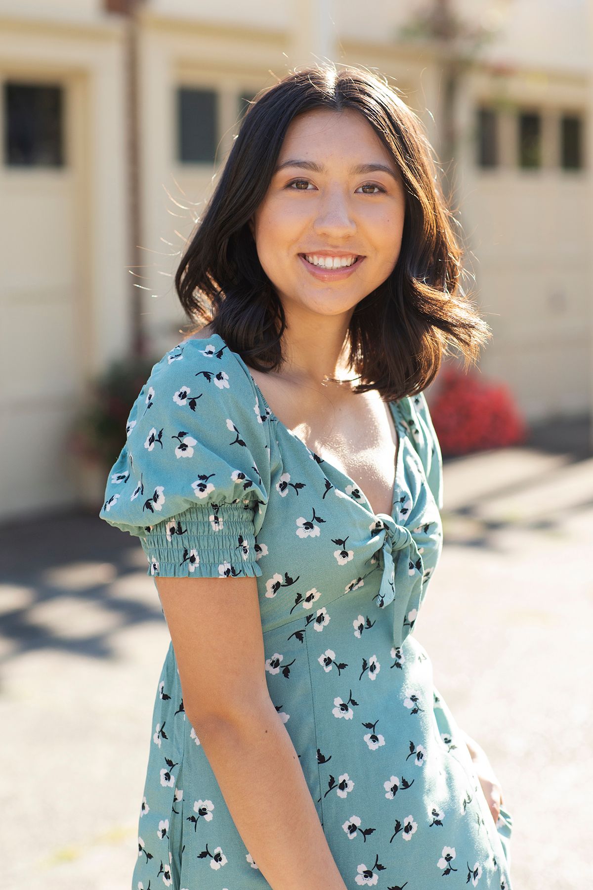 Young woman smiling outdoors in a teal floral dress, posing in front of a building on a sunny day.