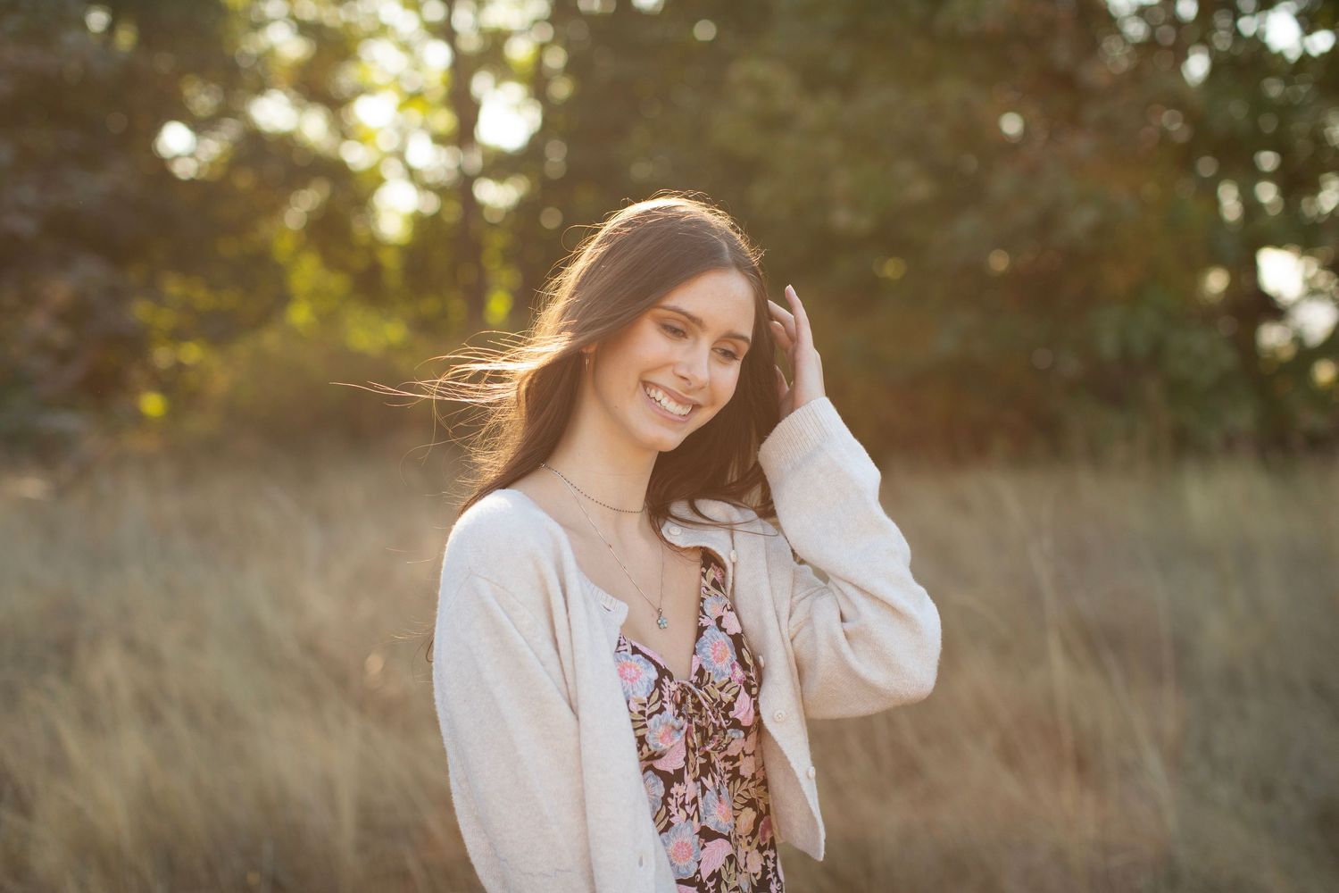 Smiling brunette in cream cardigan and floral dress poses in sunlit golden grass field outdoors.