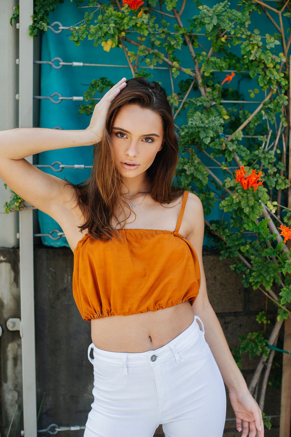 Young woman in orange crop top and white pants posing outdoors with greenery and flowers in background.