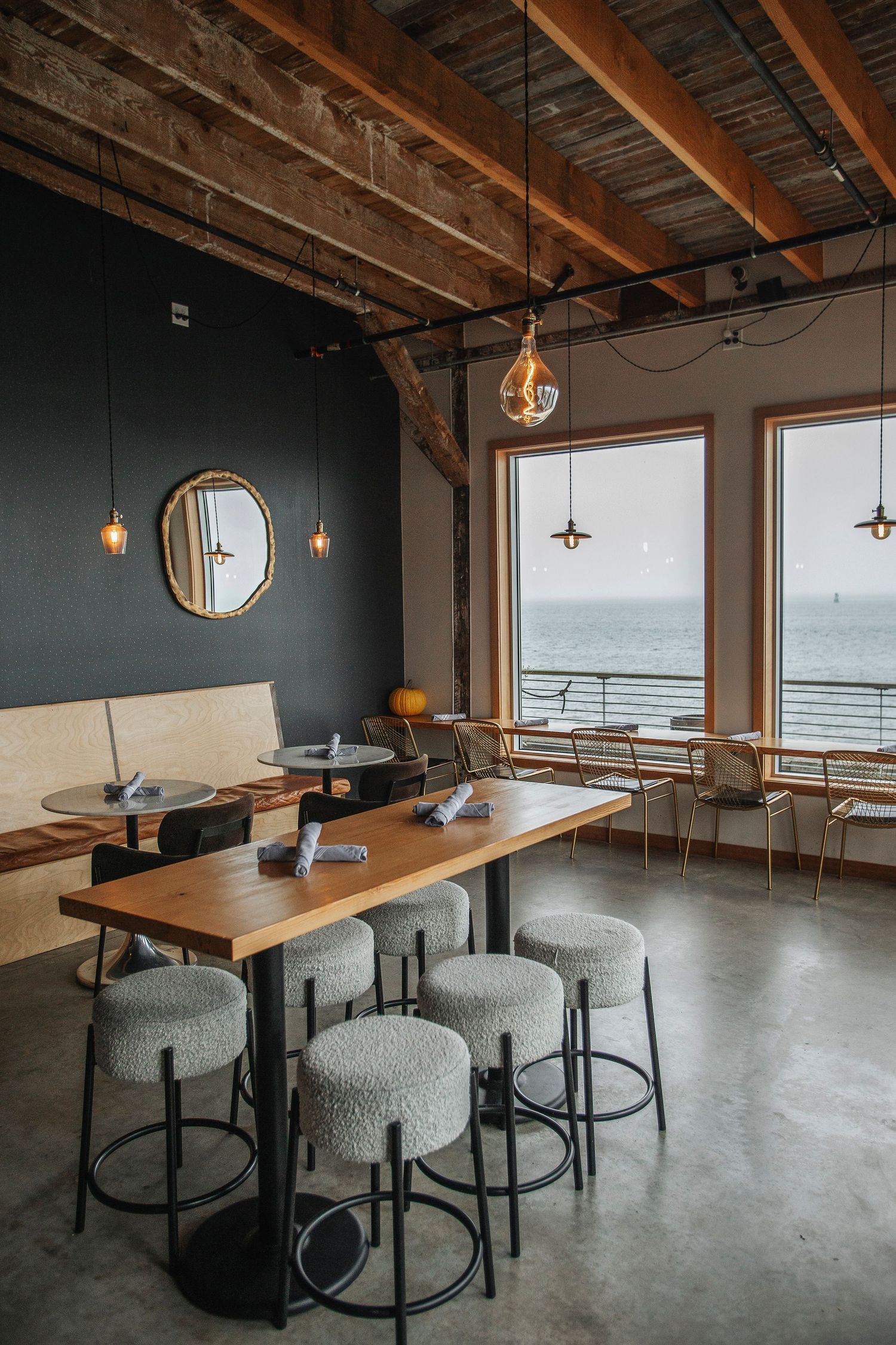 A modern restaurant interior with wooden high-top table, gray stools, exposed beam ceiling, and ocean views.