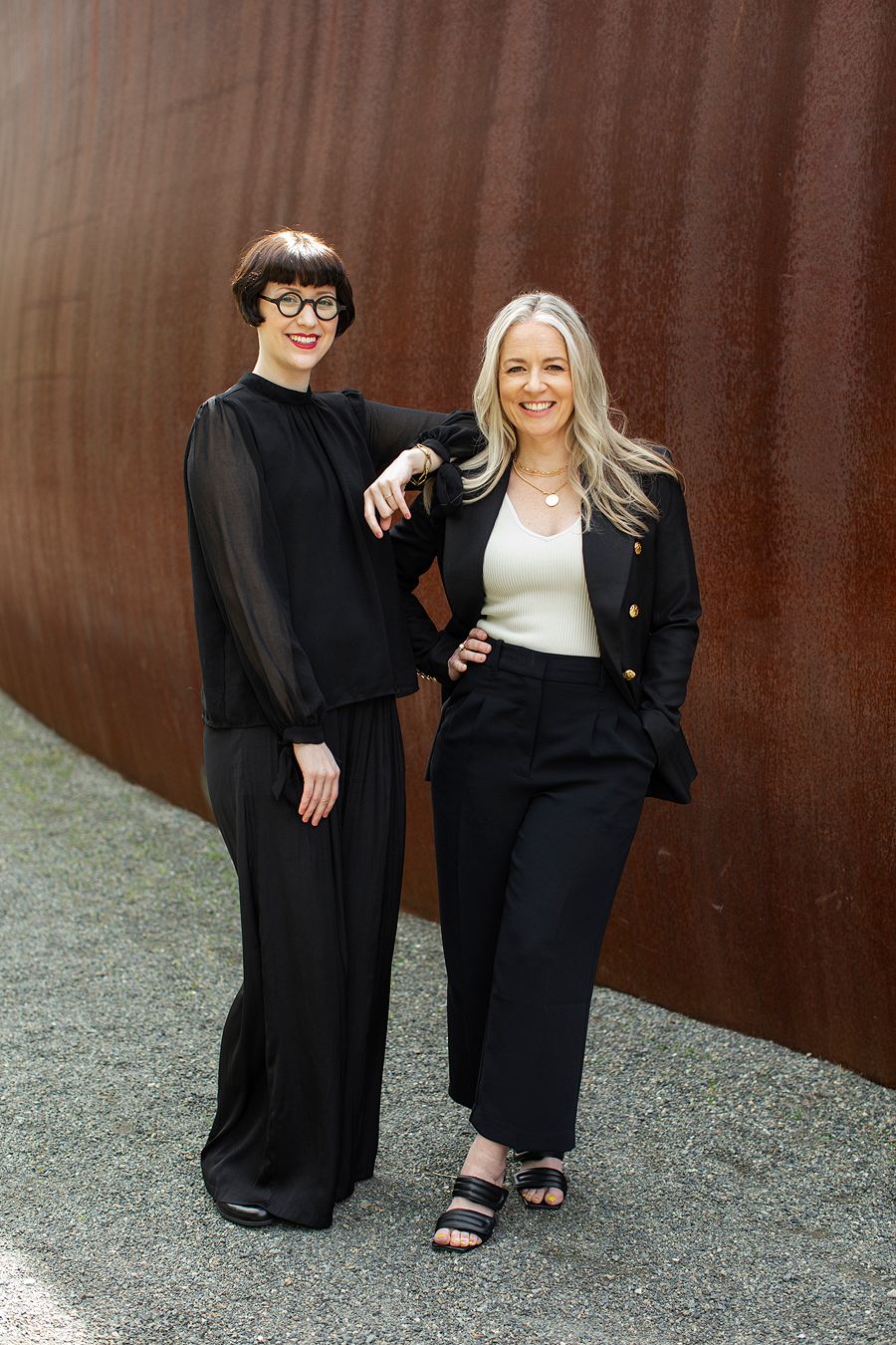 Two professional women posing together against rustic metal wall outdoors.