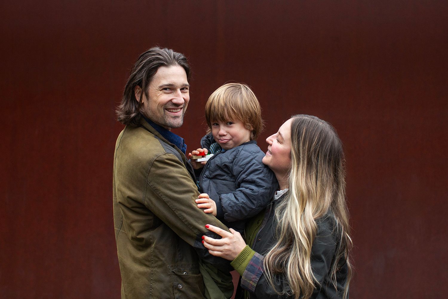 Young couple holding their toddler child while smiling together against a red wooden wall backdrop.