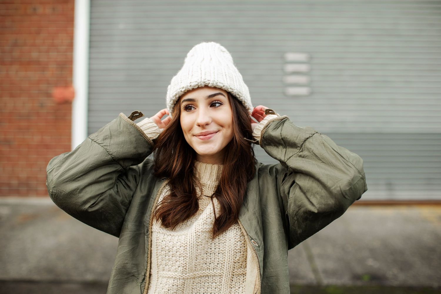 Young woman smiling in white knit beanie hat and olive green jacket outdoors in urban setting.