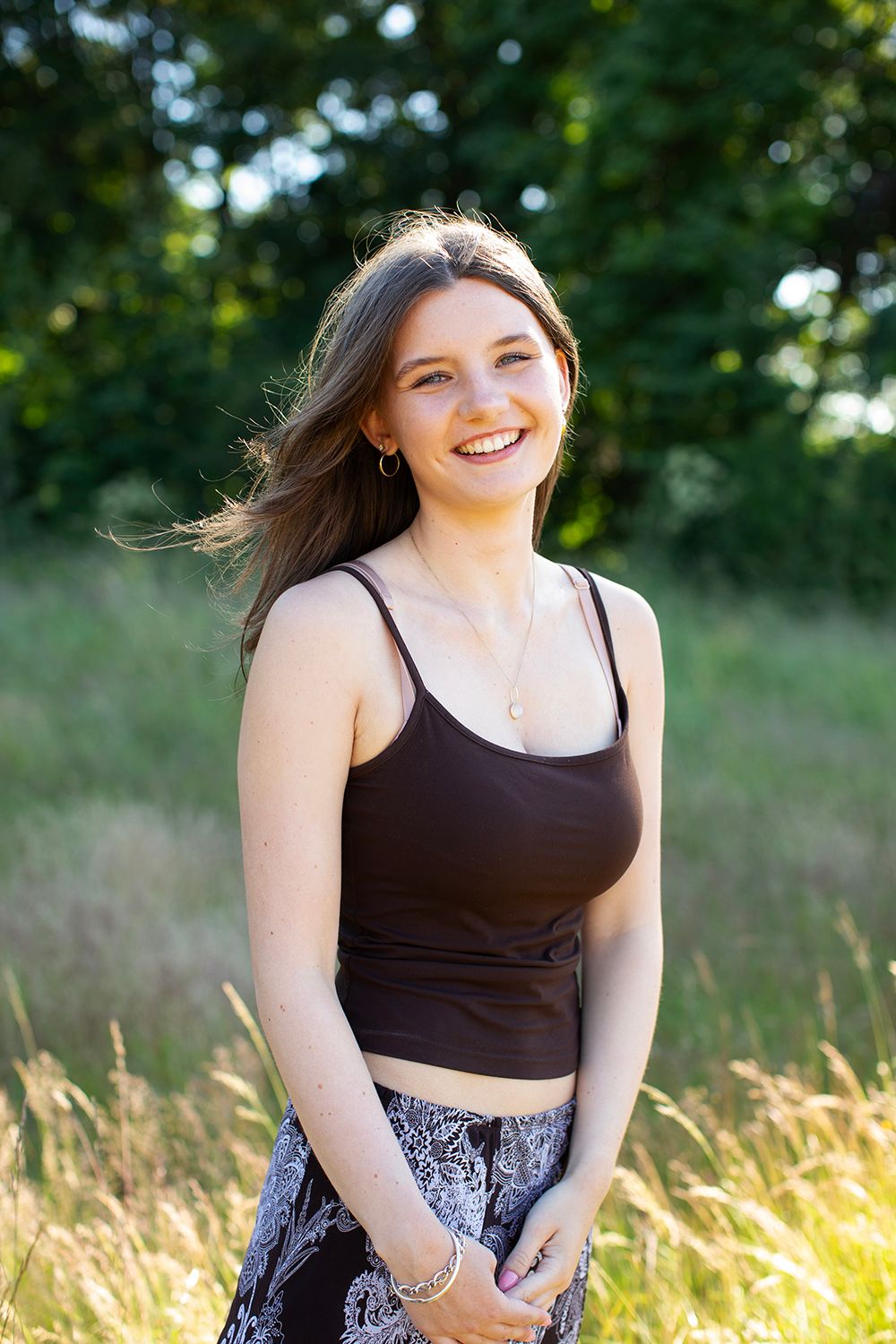 Young woman smiling outdoors in a brown crop top and floral skirt, standing in a sunlit grassy field with trees behind her.