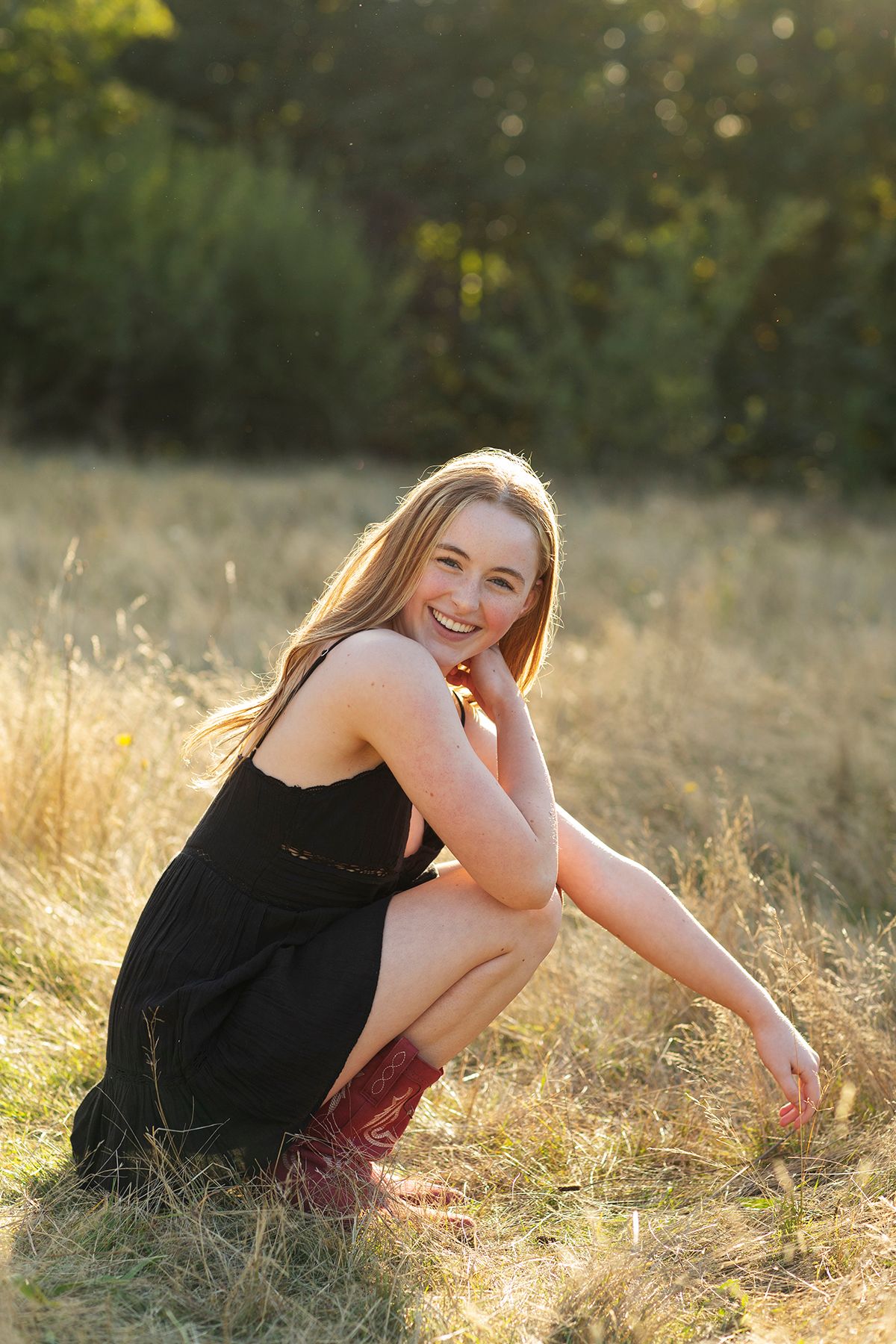 Young woman in black dress and red boots crouching in golden field, smiling brightly in warm sunset light.