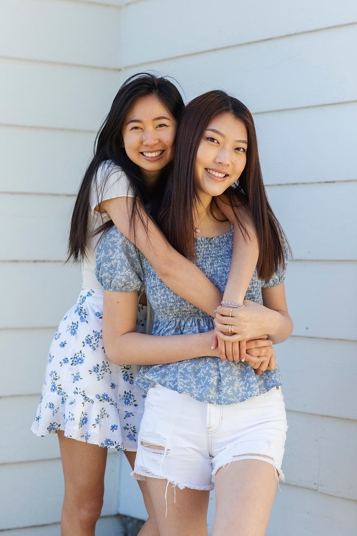 Two young Asian women smiling and hugging outdoors, wearing floral tops and white shorts against a light wall.