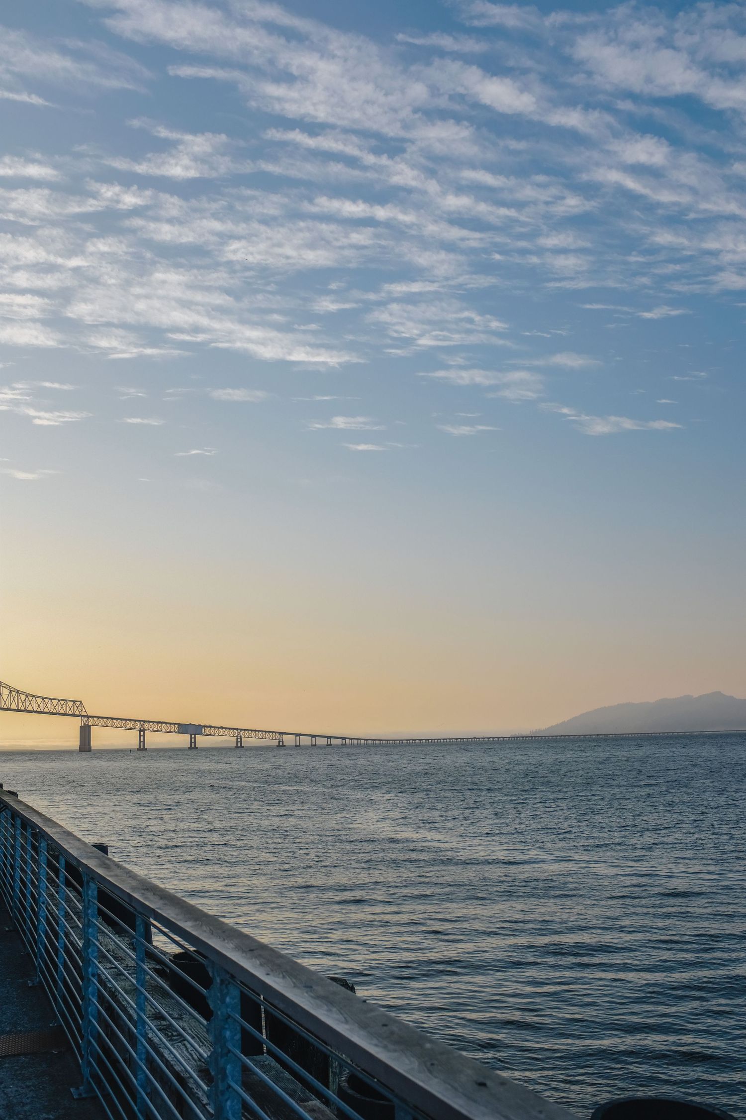 Scenic waterfront view of a long bridge stretching across calm bay waters at golden hour with wispy clouds above.