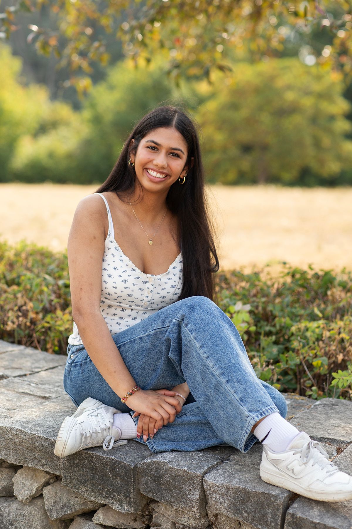 Smiling young woman in floral tank top and jeans sitting on stone wall with autumn trees behind her.