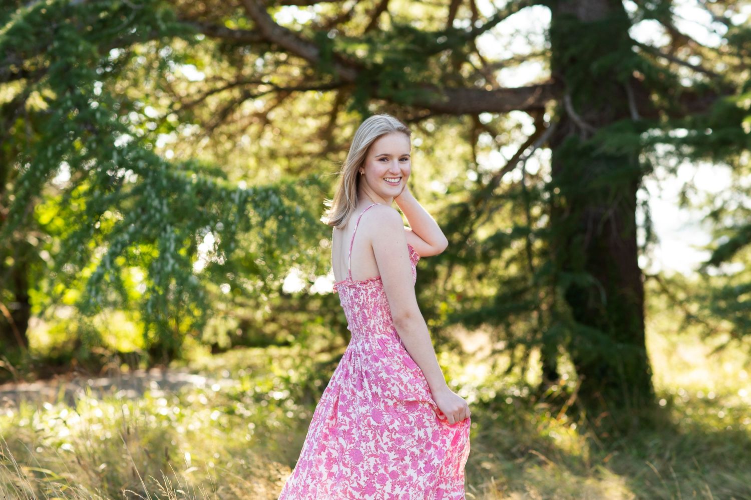 Smiling young woman in pink floral dress posing outdoors near evergreen trees.