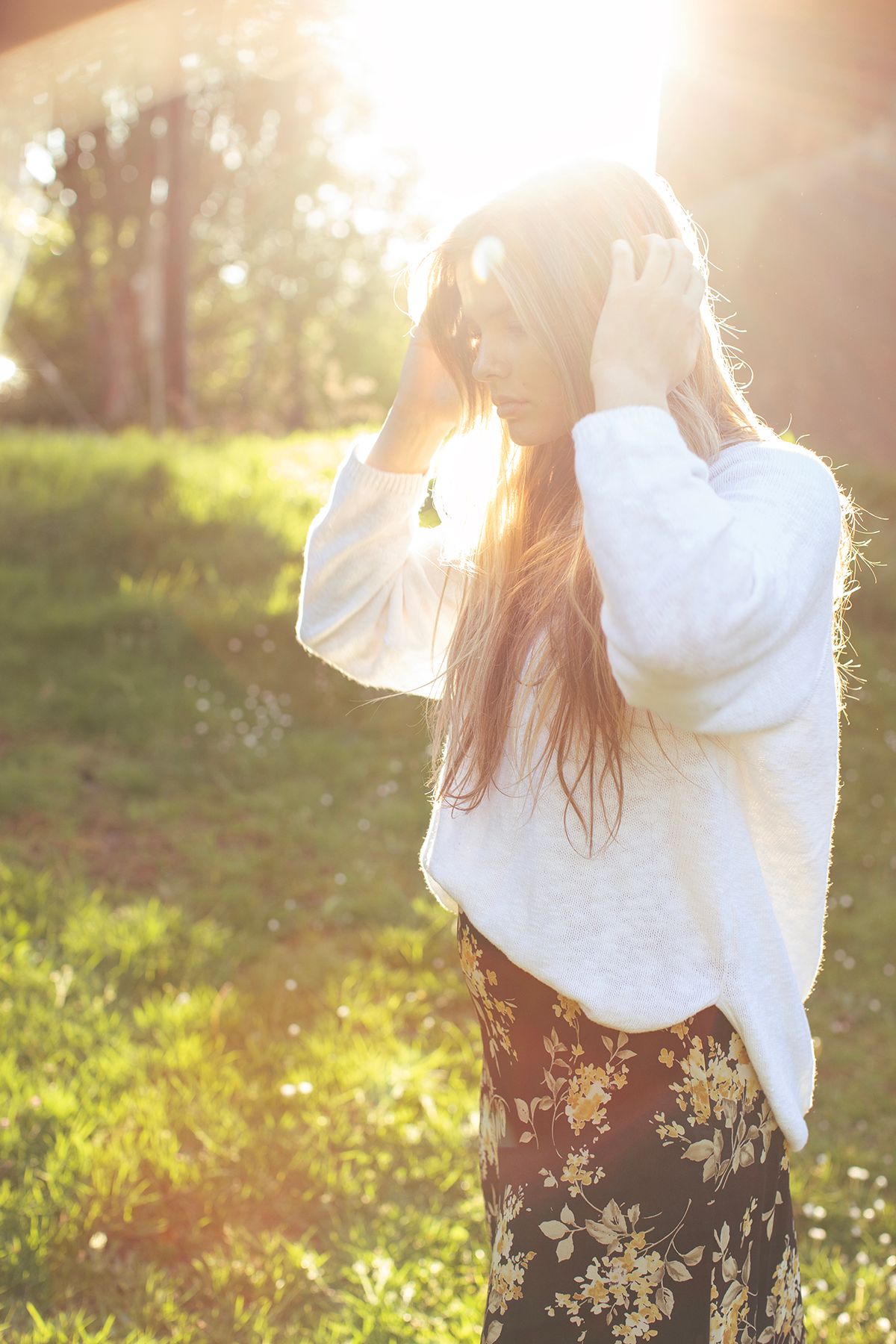 Woman in white sweater and floral skirt standing in sunlit park with long hair blowing in the breeze.