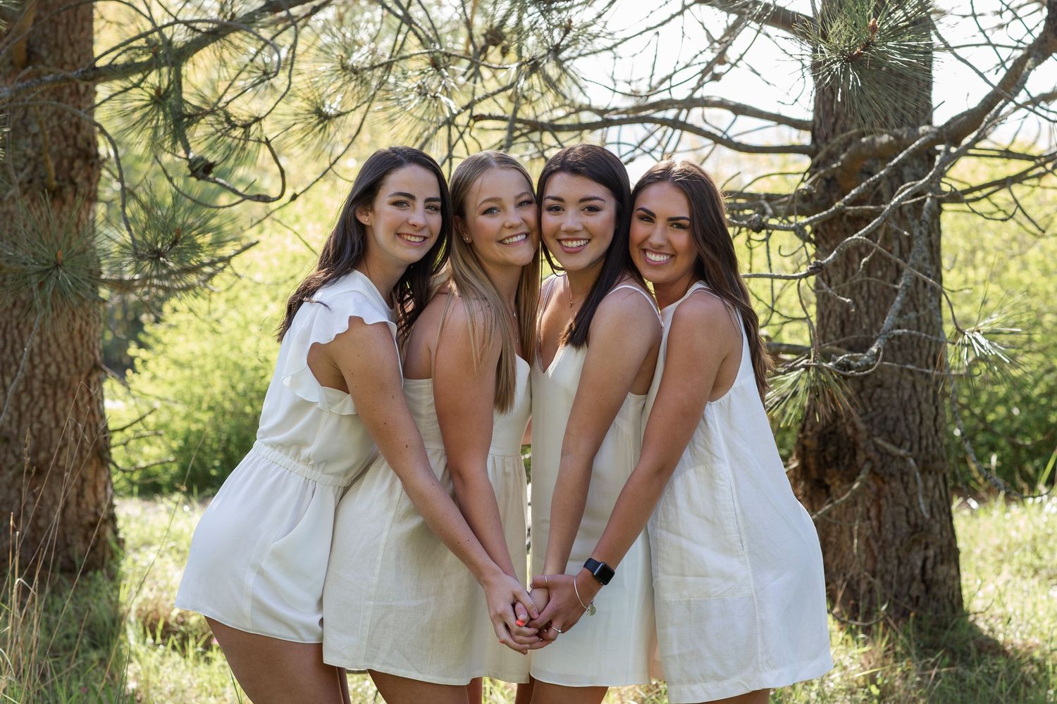 Four young women in white dresses smiling and posing together outdoors under pine trees on a sunny day.