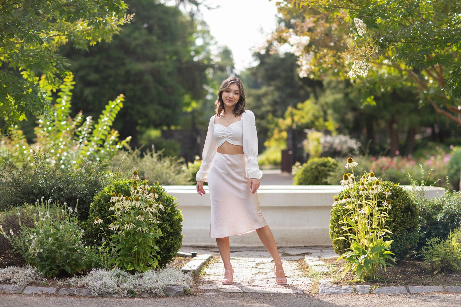 Young woman in a white crop top and blush midi skirt posing barefoot by a garden stone fountain.
