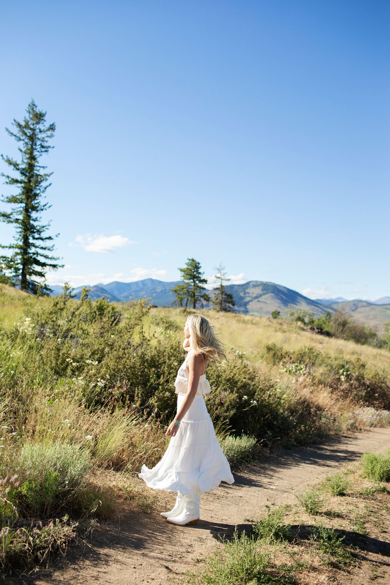 Blonde woman in flowing white dress walking on a scenic mountain trail under blue sky.
