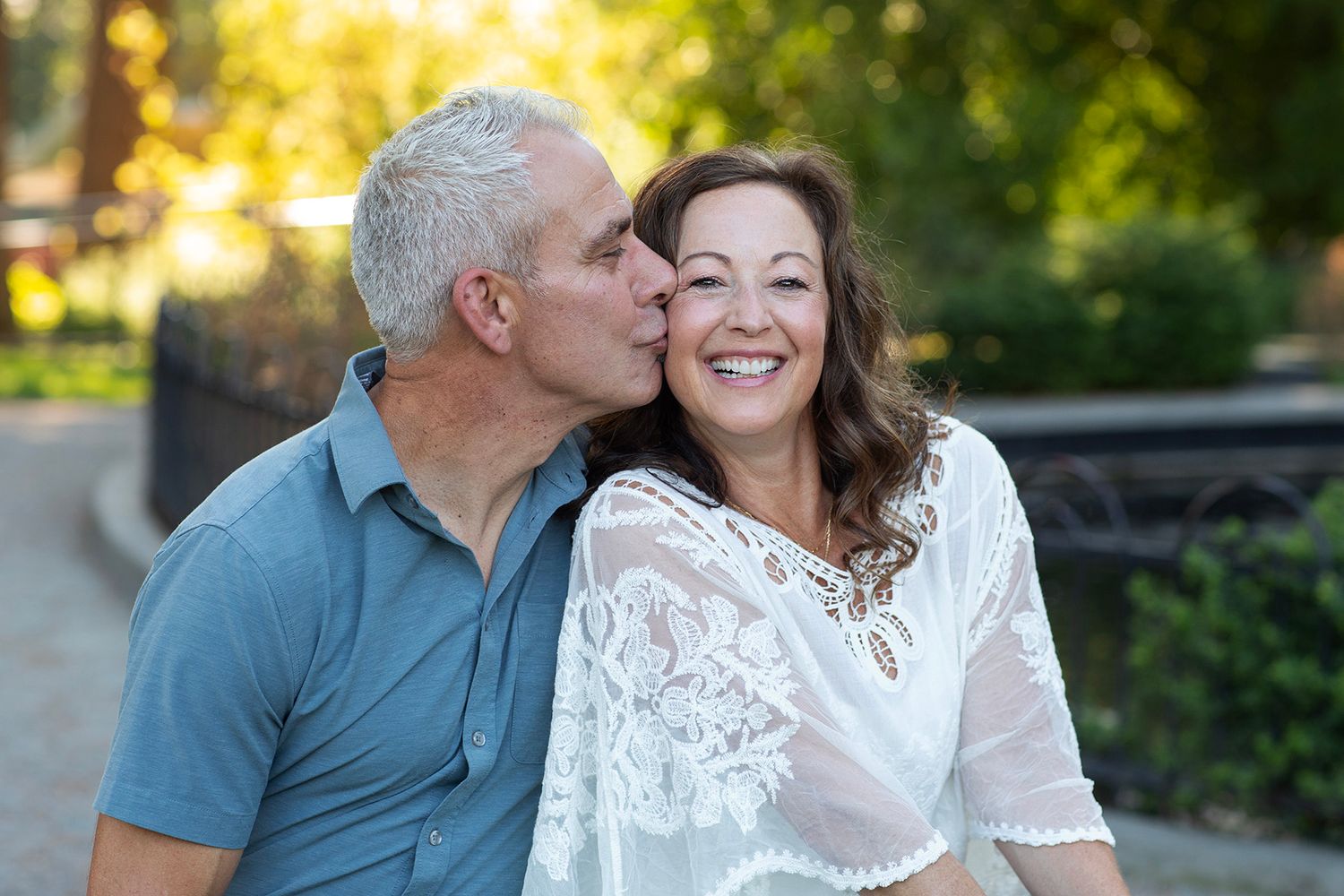 A smiling middle-aged couple sharing a romantic kiss outdoors in a sunny park setting.