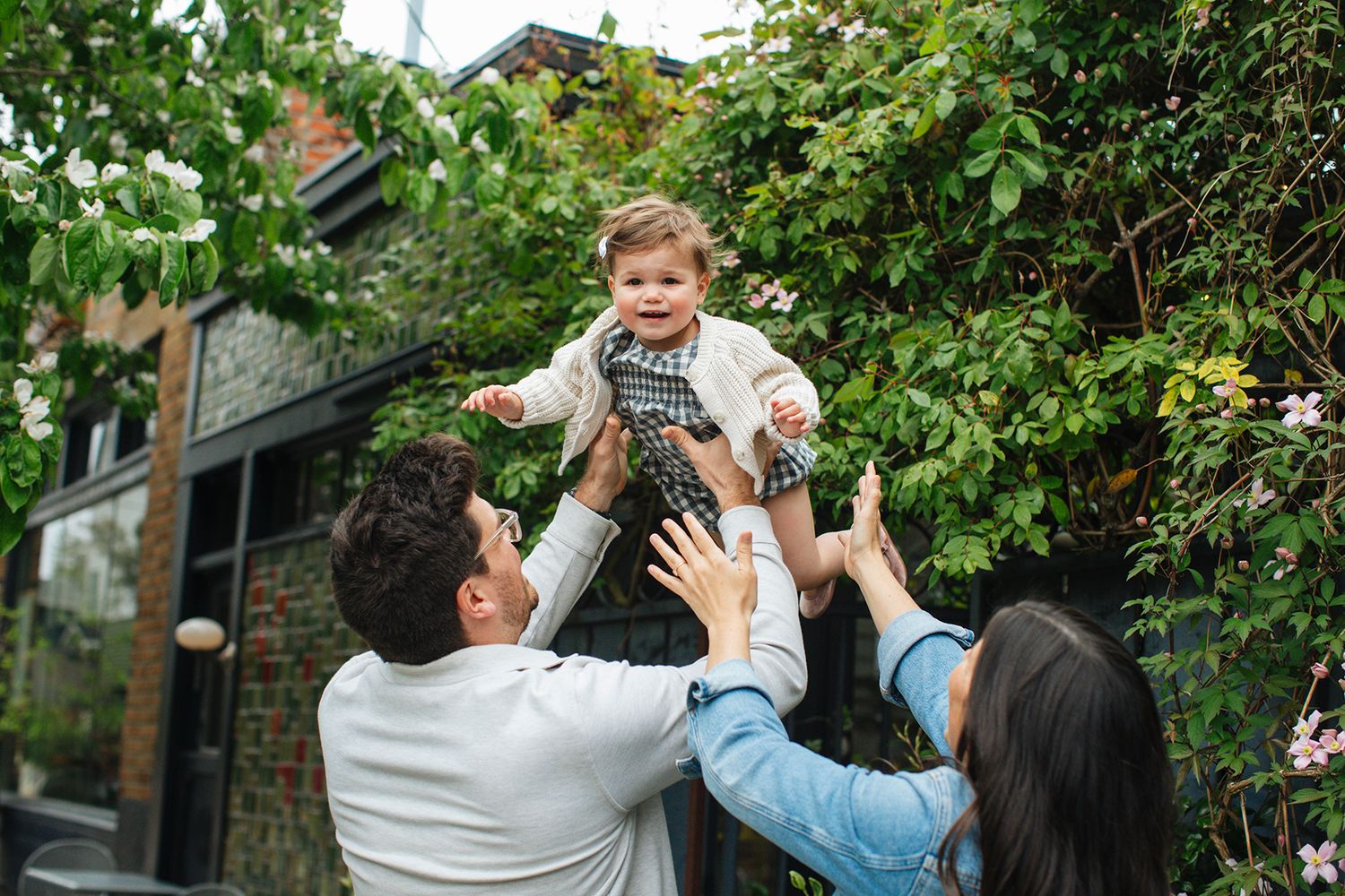 Happy family with parents lifting smiling baby outdoors in lush garden near brick house on a sunny day.