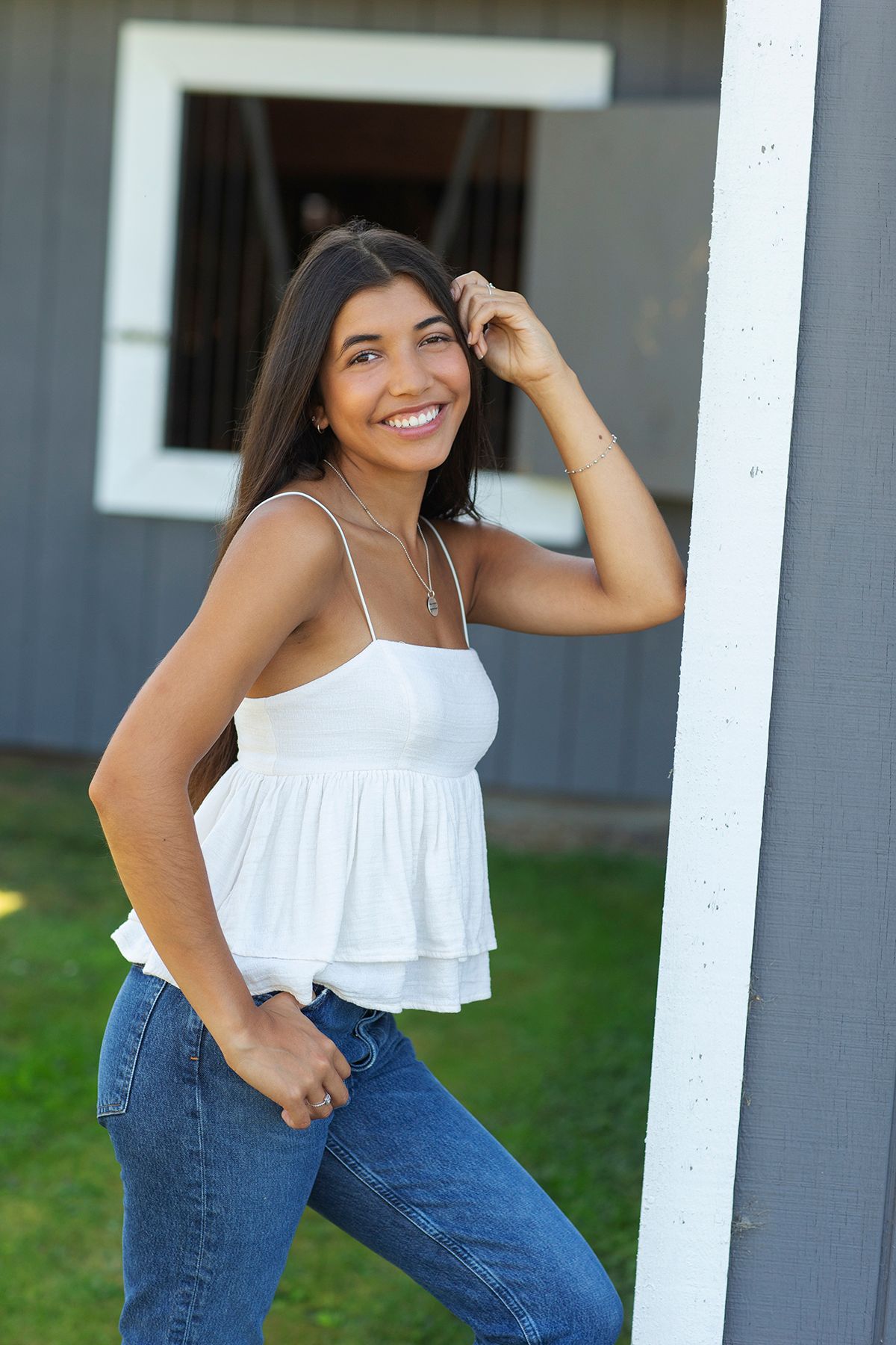 Smiling young woman in white camisole top and jeans posing outdoors near a white post with a barn in the background.