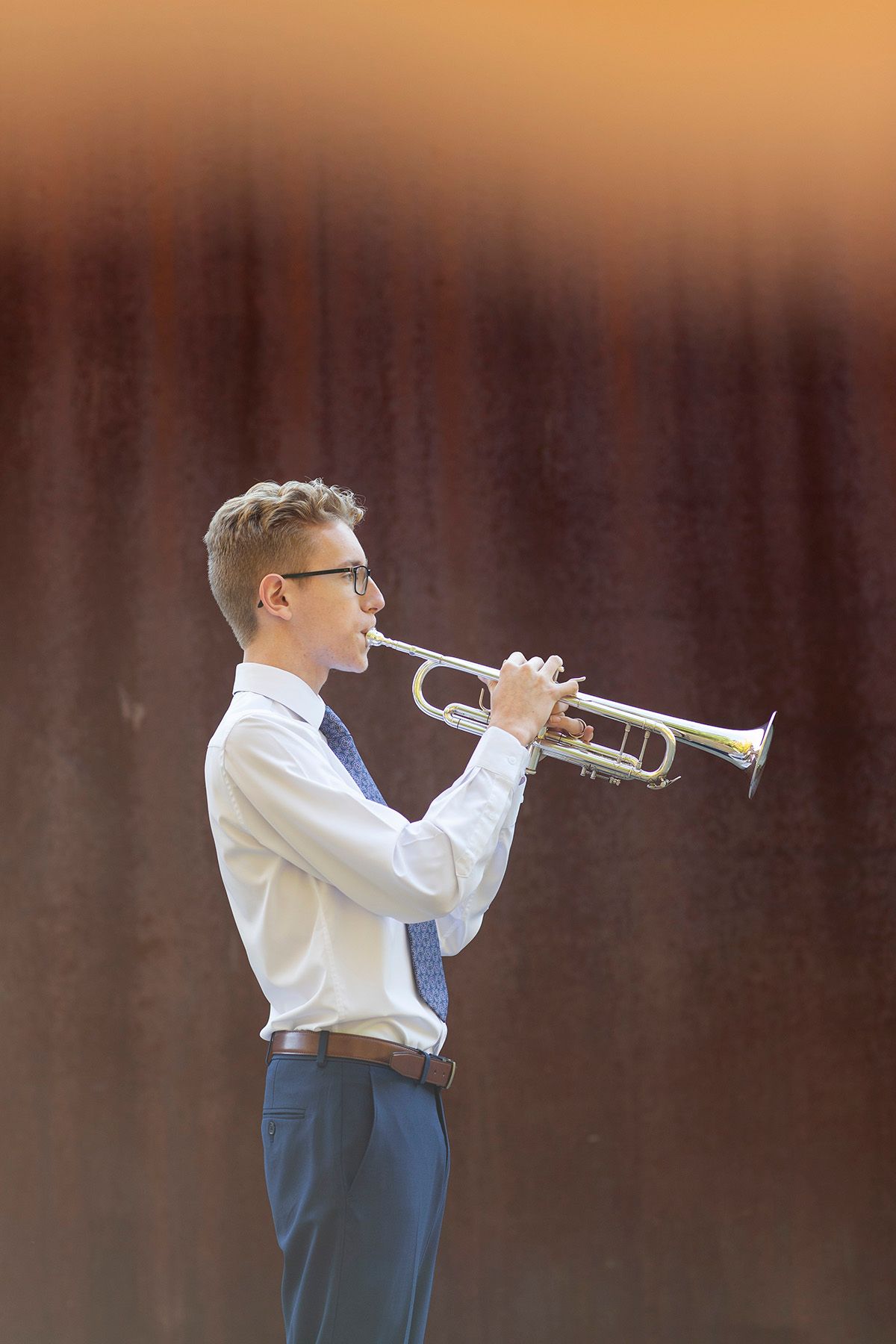 Young man in white shirt and blue pants playing trumpet outdoors against warm orange backdrop.