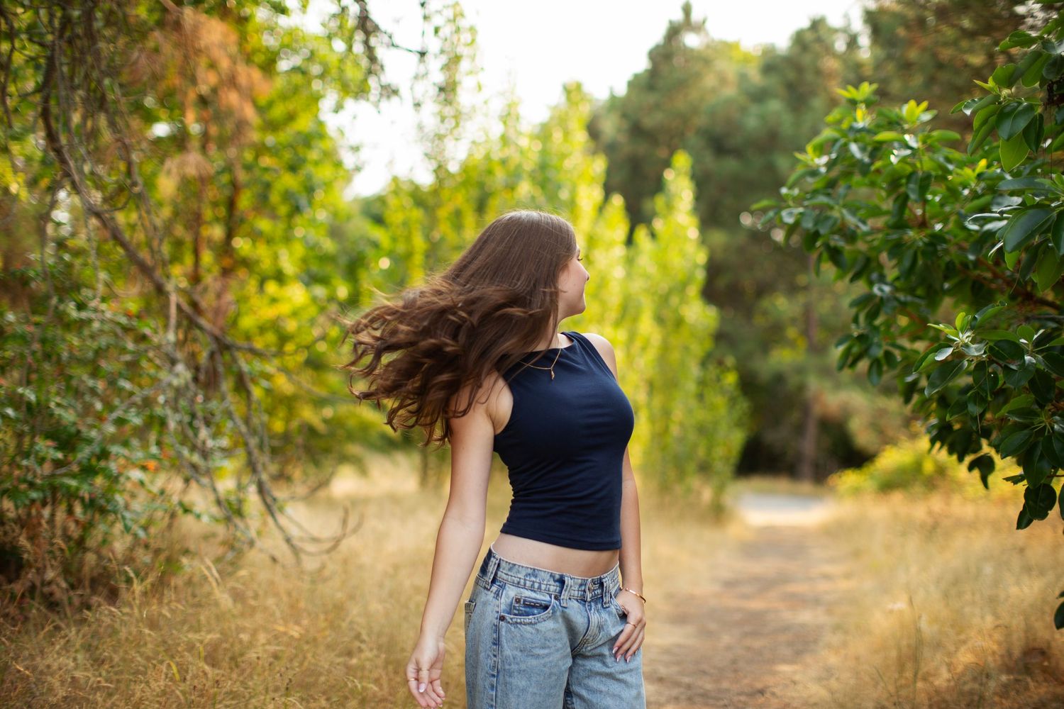 Young woman in crop top and jeans tossing her long hair on a scenic outdoor path surrounded by green trees.