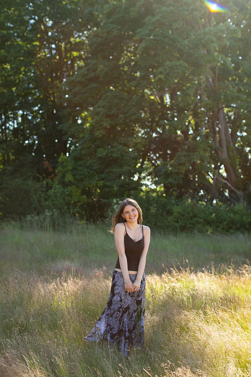 Young woman smiling in black top and floral skirt standing in sunlit green meadow with trees.
