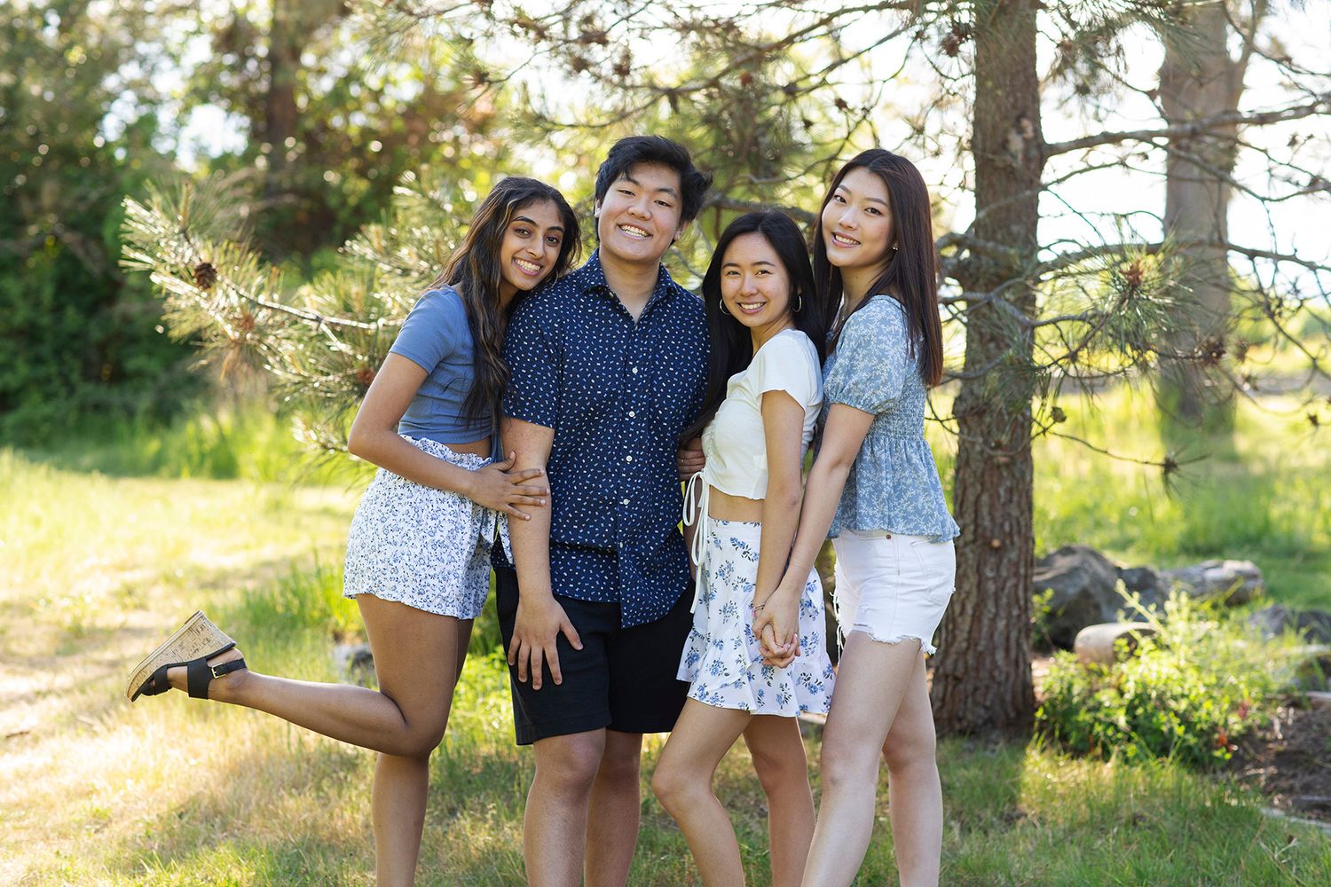 Four smiling young adults posing together outdoors in a sunny park surrounded by green trees and natural scenery.