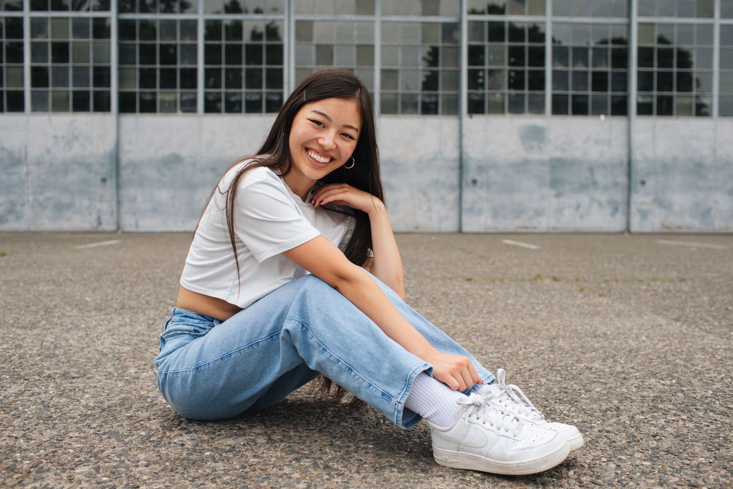 Smiling young woman sitting on ground outdoors wearing white crop top, blue jeans, and white sneakers.