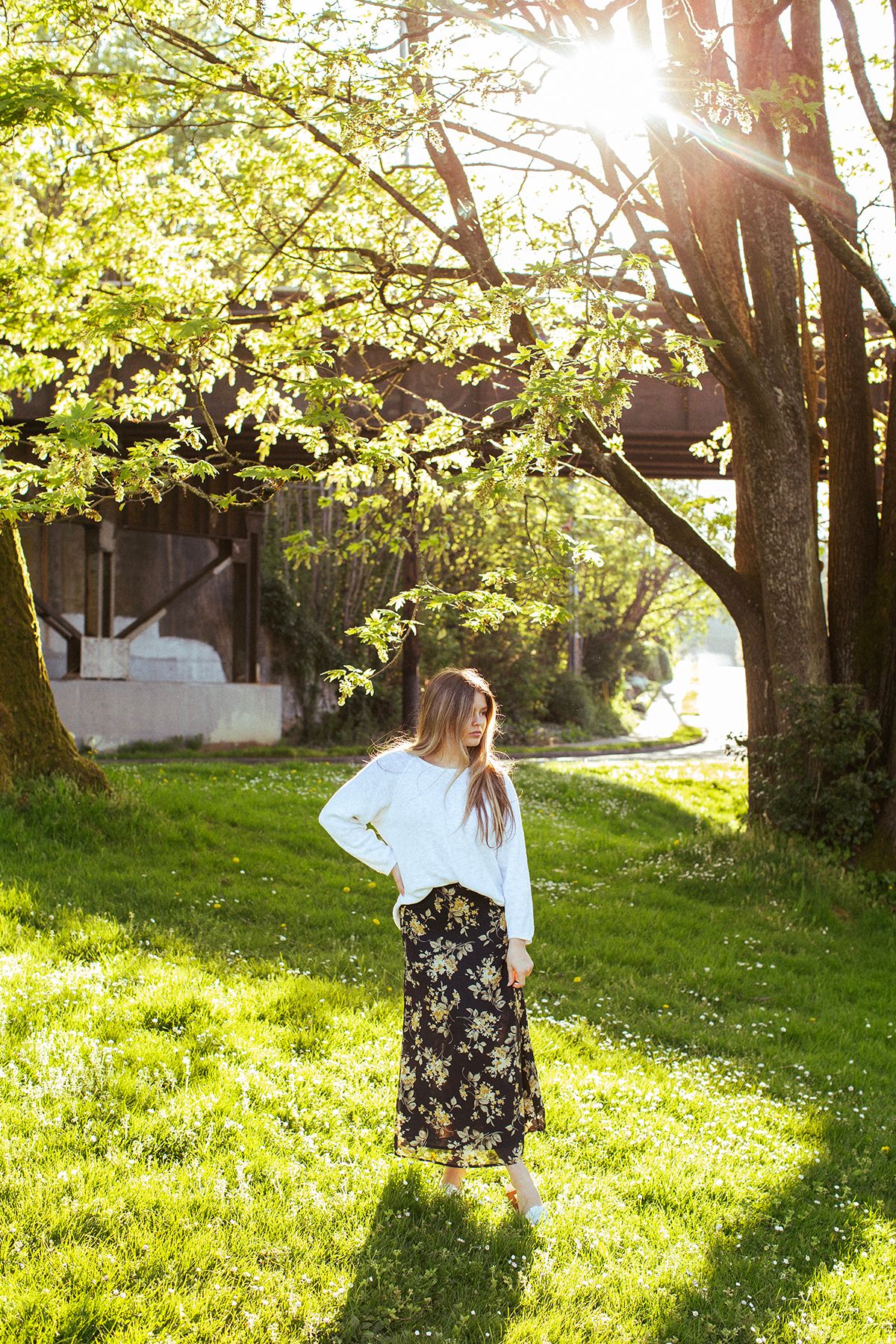 Young woman in white top and floral skirt standing on green grass under sunlit trees near a rustic building.