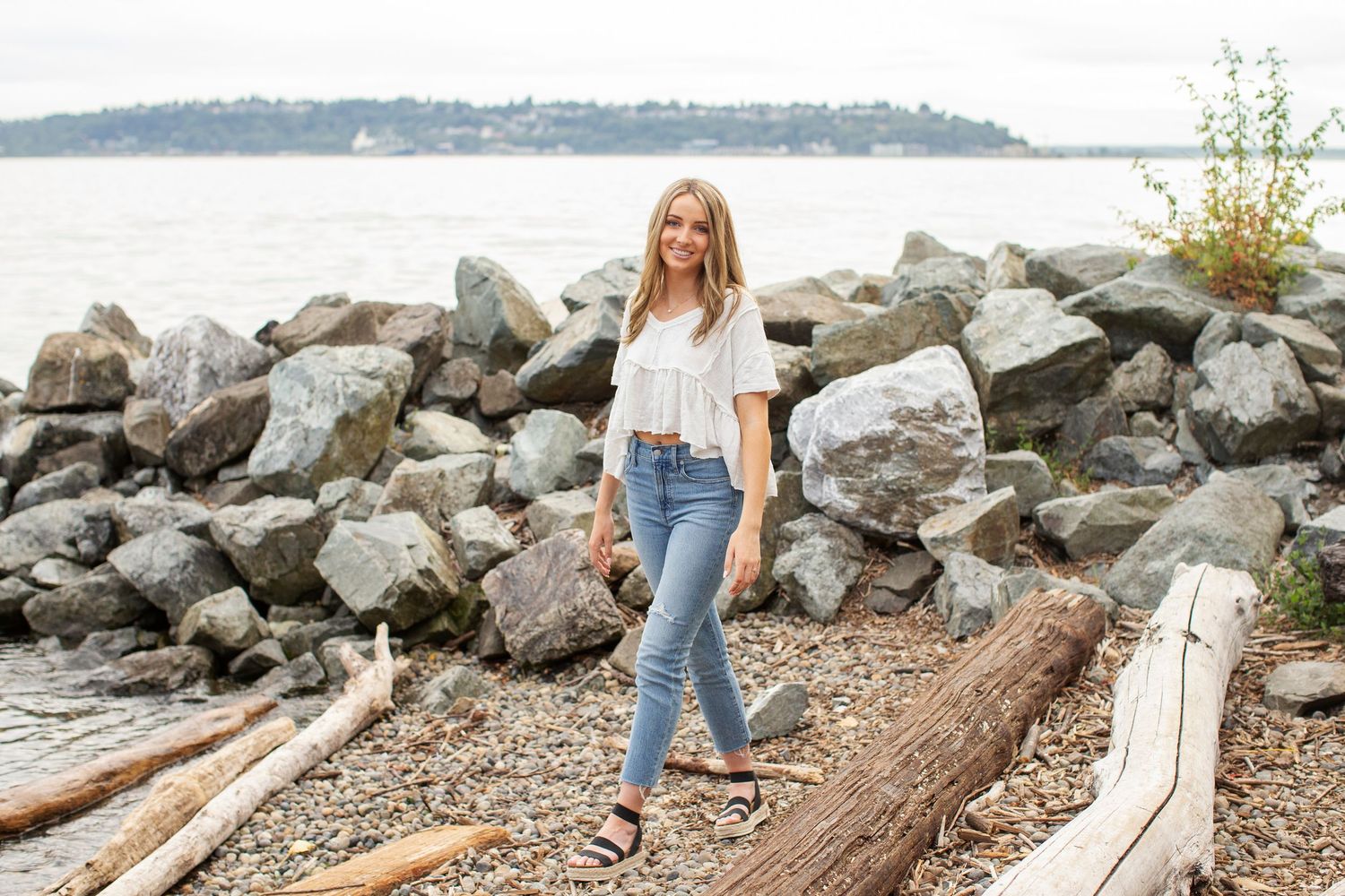 Young woman in white top and jeans standing on rocky beach near waterfront.