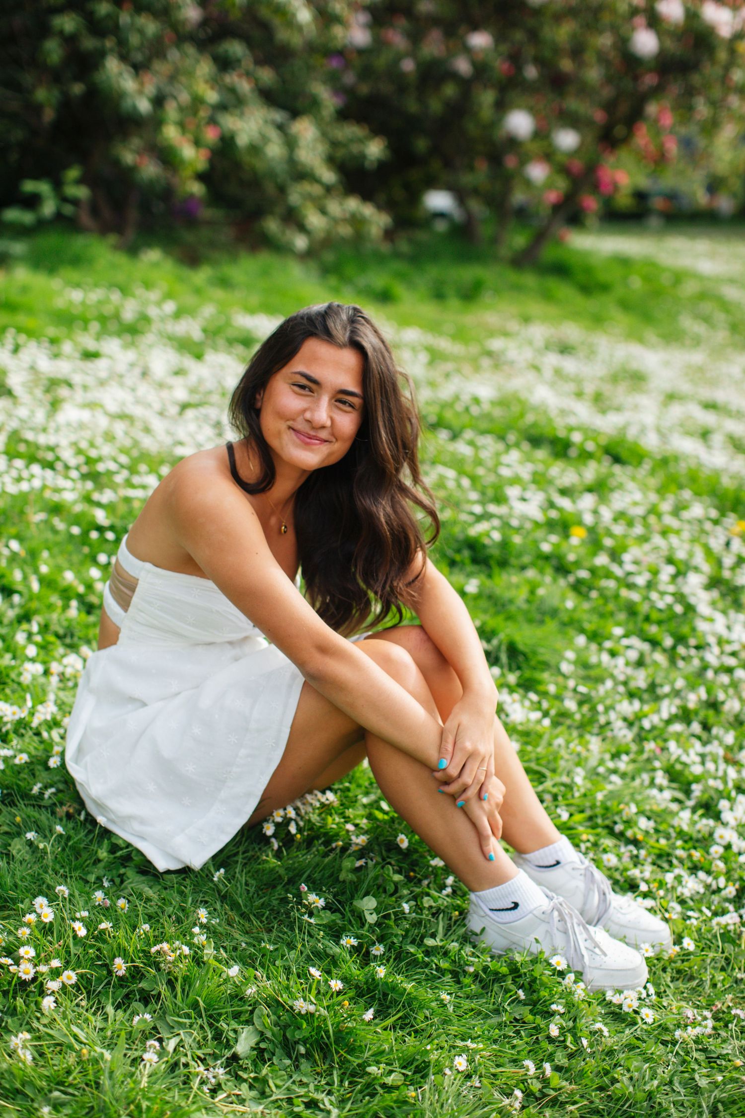 Young woman in white dress and sneakers sitting on grass field with wildflowers, smiling at camera on sunny day.