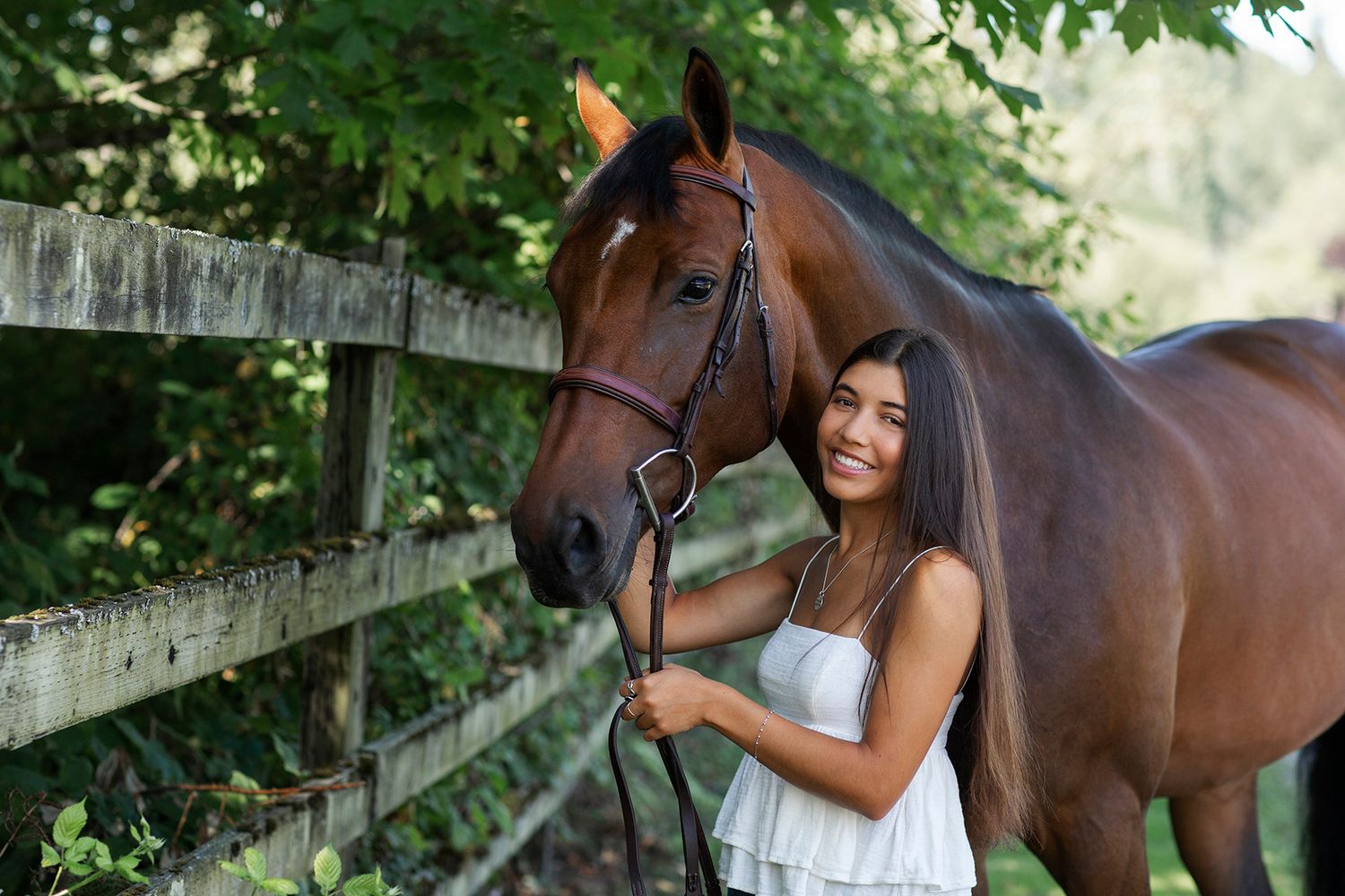 Smiling young woman in white dress standing beside a brown horse near a wooden fence surrounded by lush greenery.