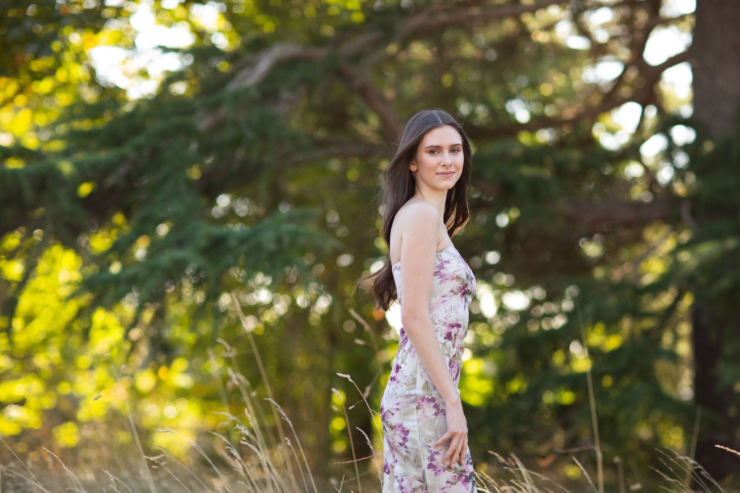 Brunette woman in floral maxi dress walks through golden grass field surrounded by tall green trees.
