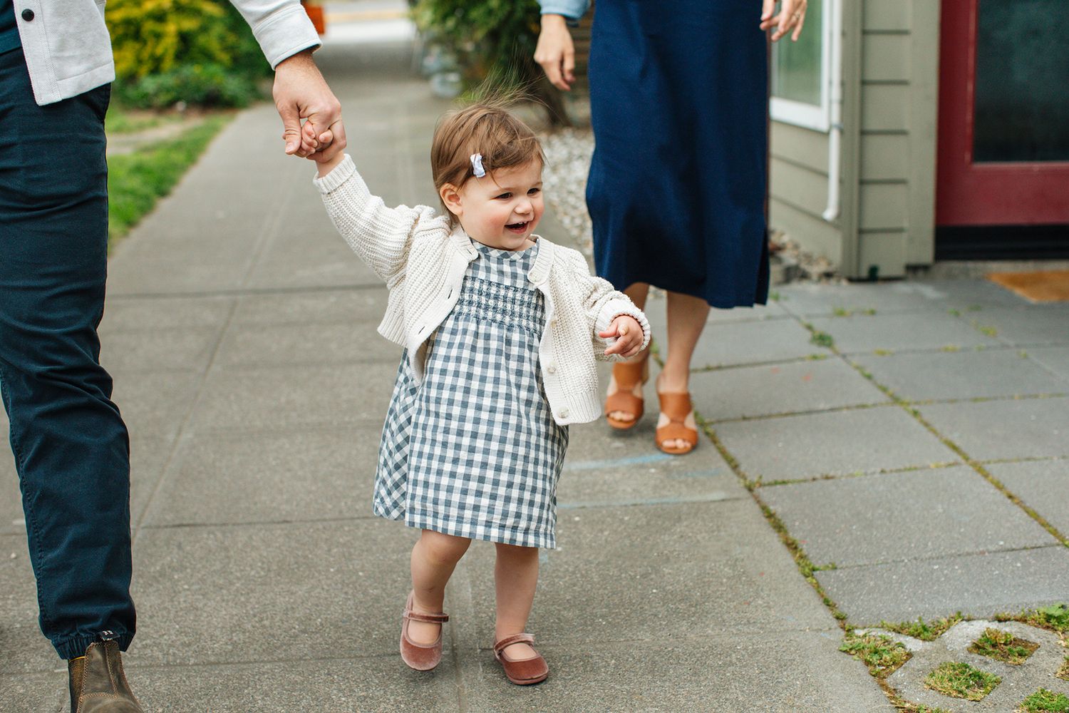 Toddler girl in gingham dress and white jacket walks holding parents hands on a sidewalk.