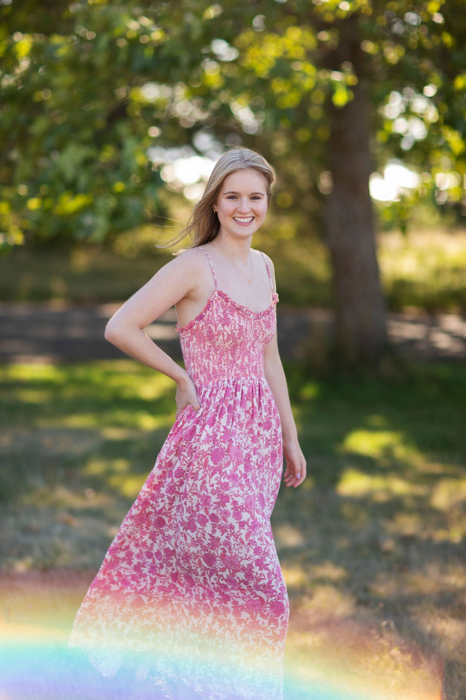 Blonde woman in flowing pink floral maxi dress smiling in sunlit park with rainbow reflection.