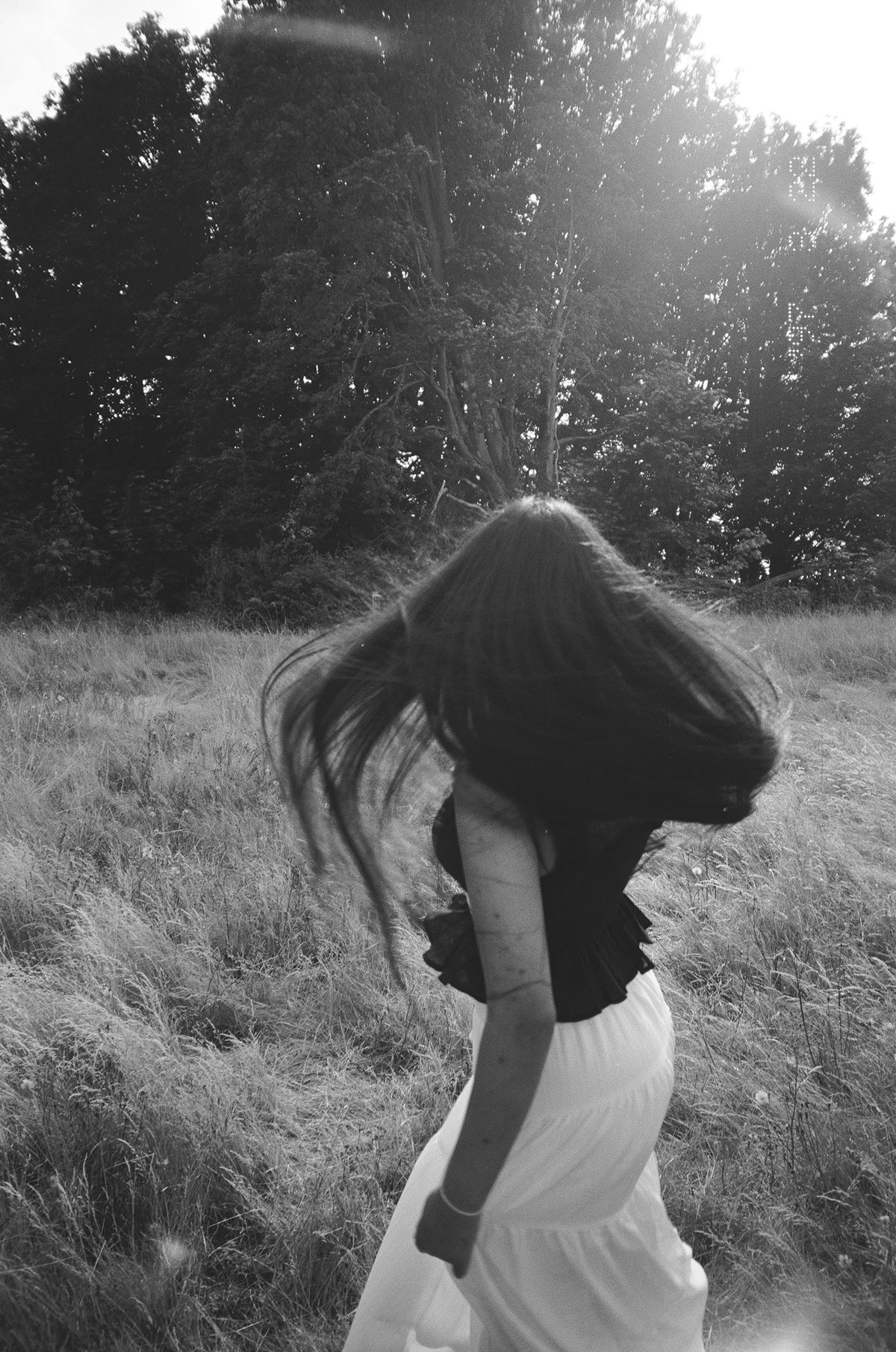 Black and white photo of woman with flowing hair twirling in white skirt in sunny field.