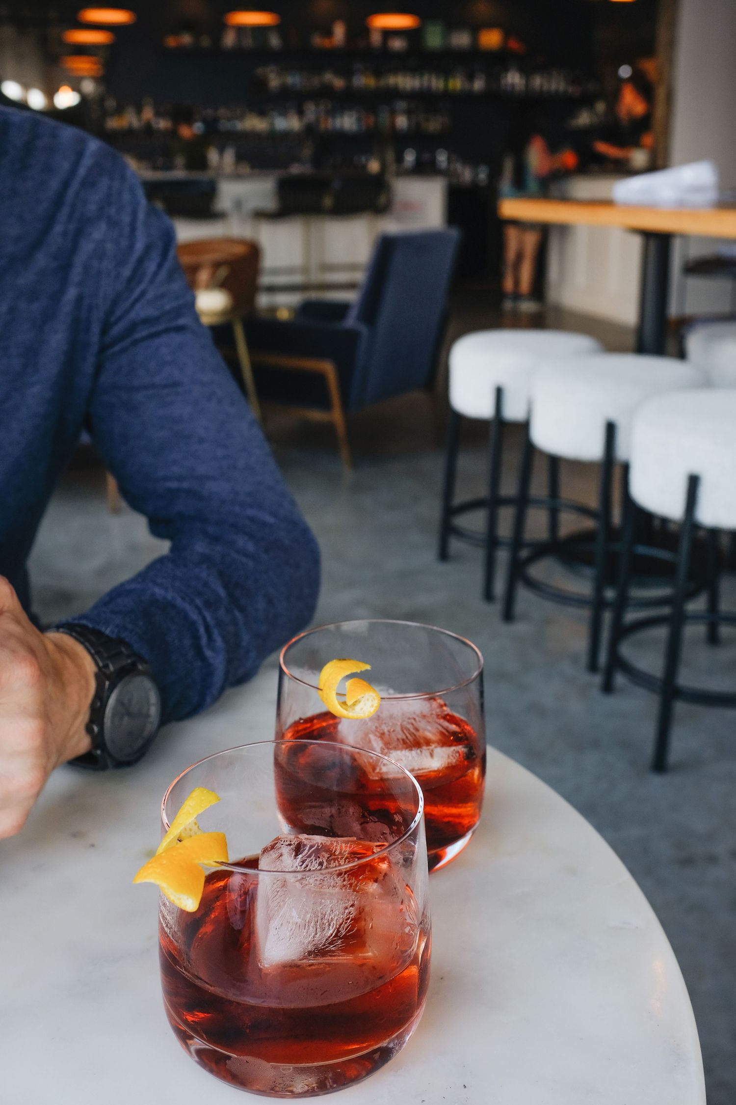 Two red Negroni cocktails with orange peel garnish on a marble table in a modern bar setting.