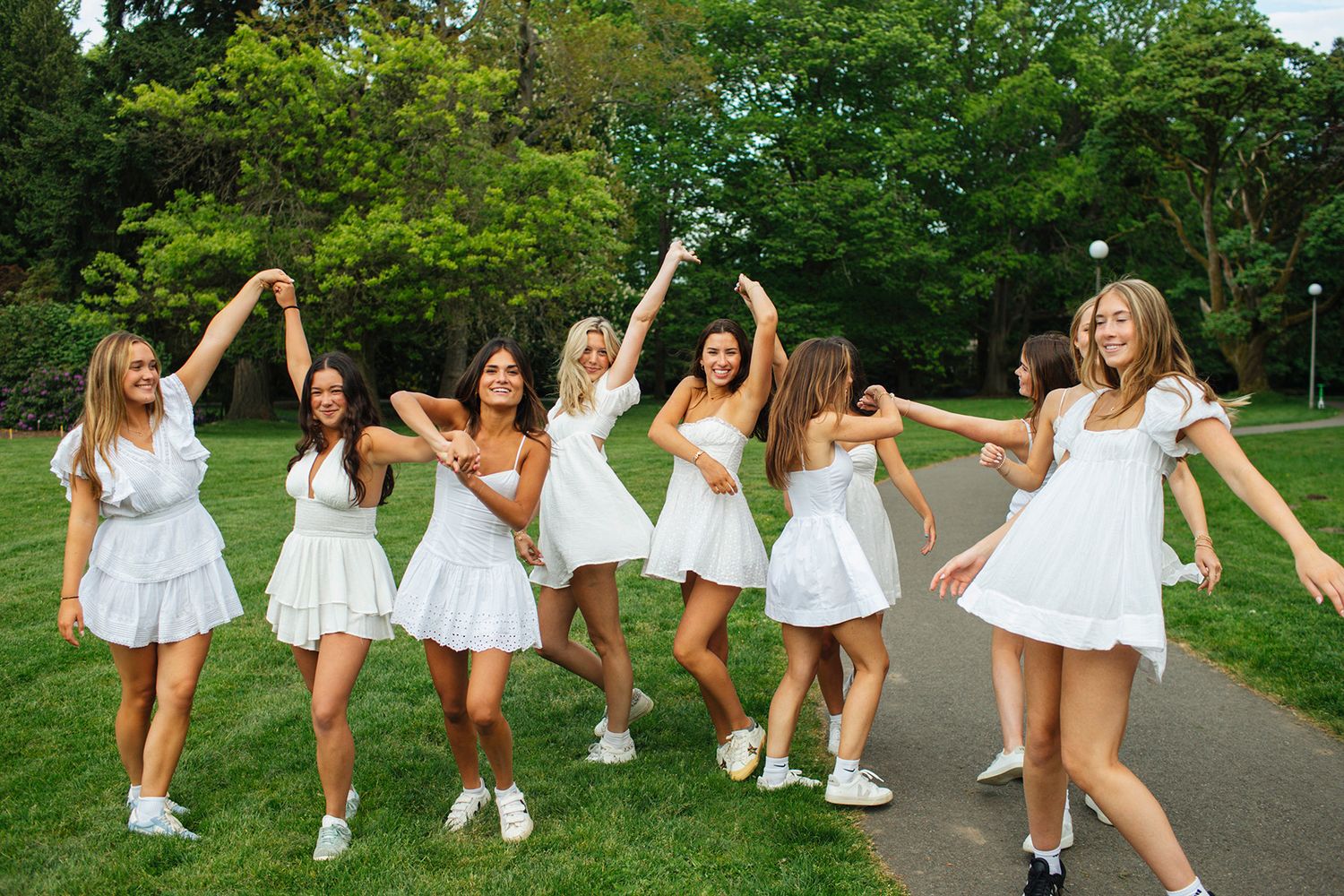 Group of eight women in white dresses dancing and celebrating together on a park pathway surrounded by green trees.