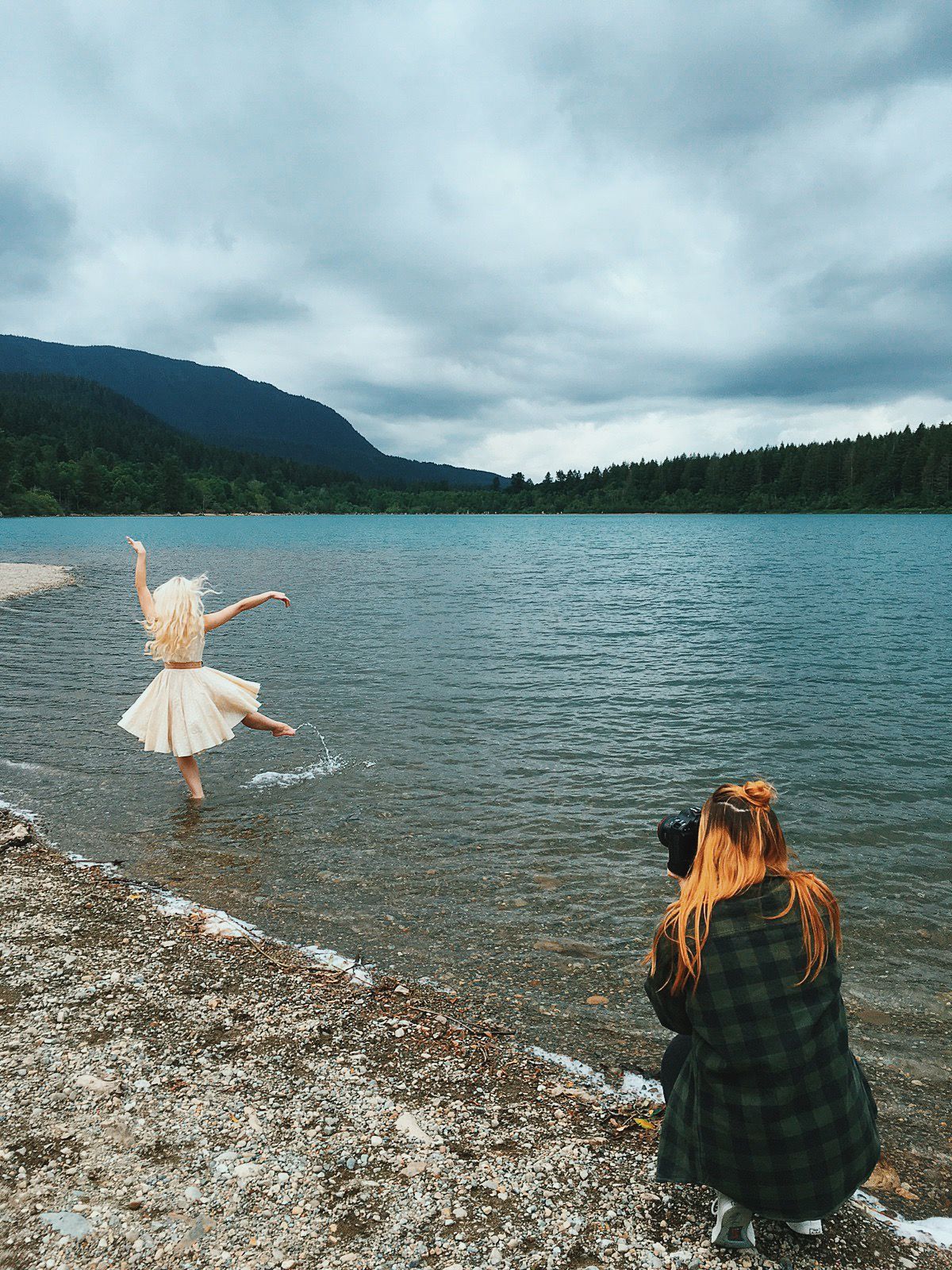 Photographer crouching on rocky lakeshore capturing a woman dancing in white dress near mountain lake.