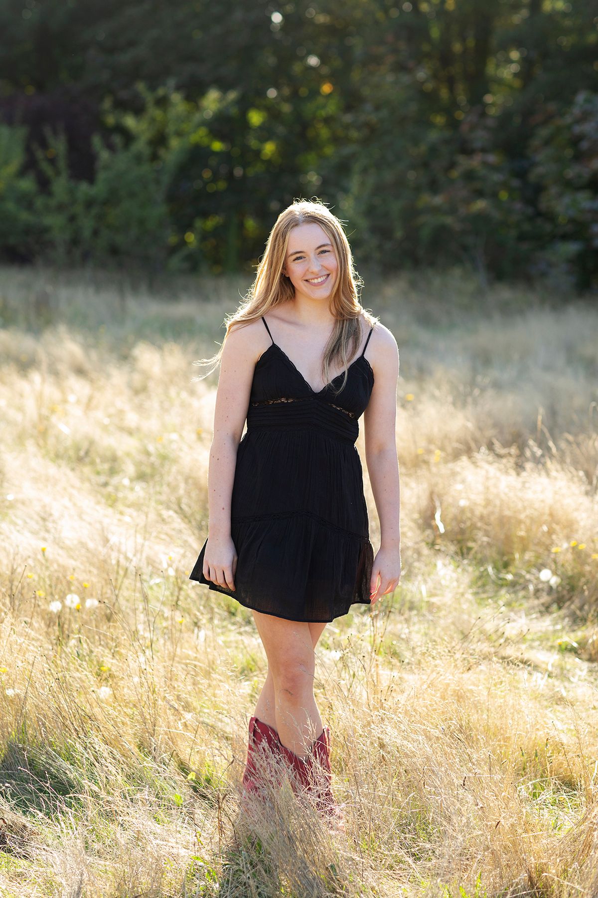 Smiling blonde woman in black sundress and red cowboy boots standing in golden field with trees behind her.