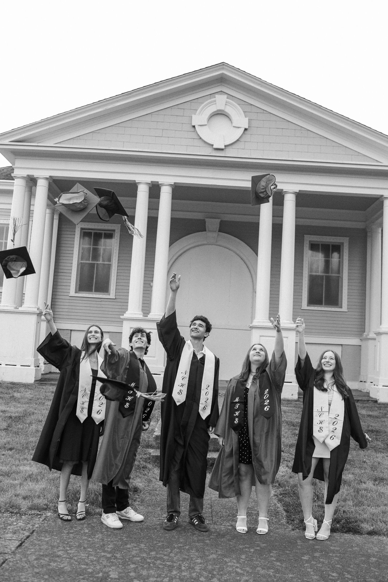 Five graduates in black caps and gowns toss their mortarboards in the air outside a classic white building.