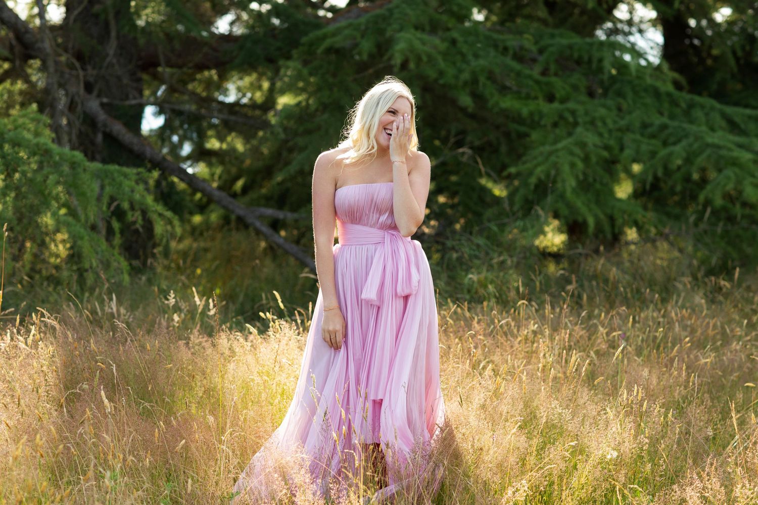 Blonde woman in a flowing pink strapless gown standing in a golden meadow under a large tree.