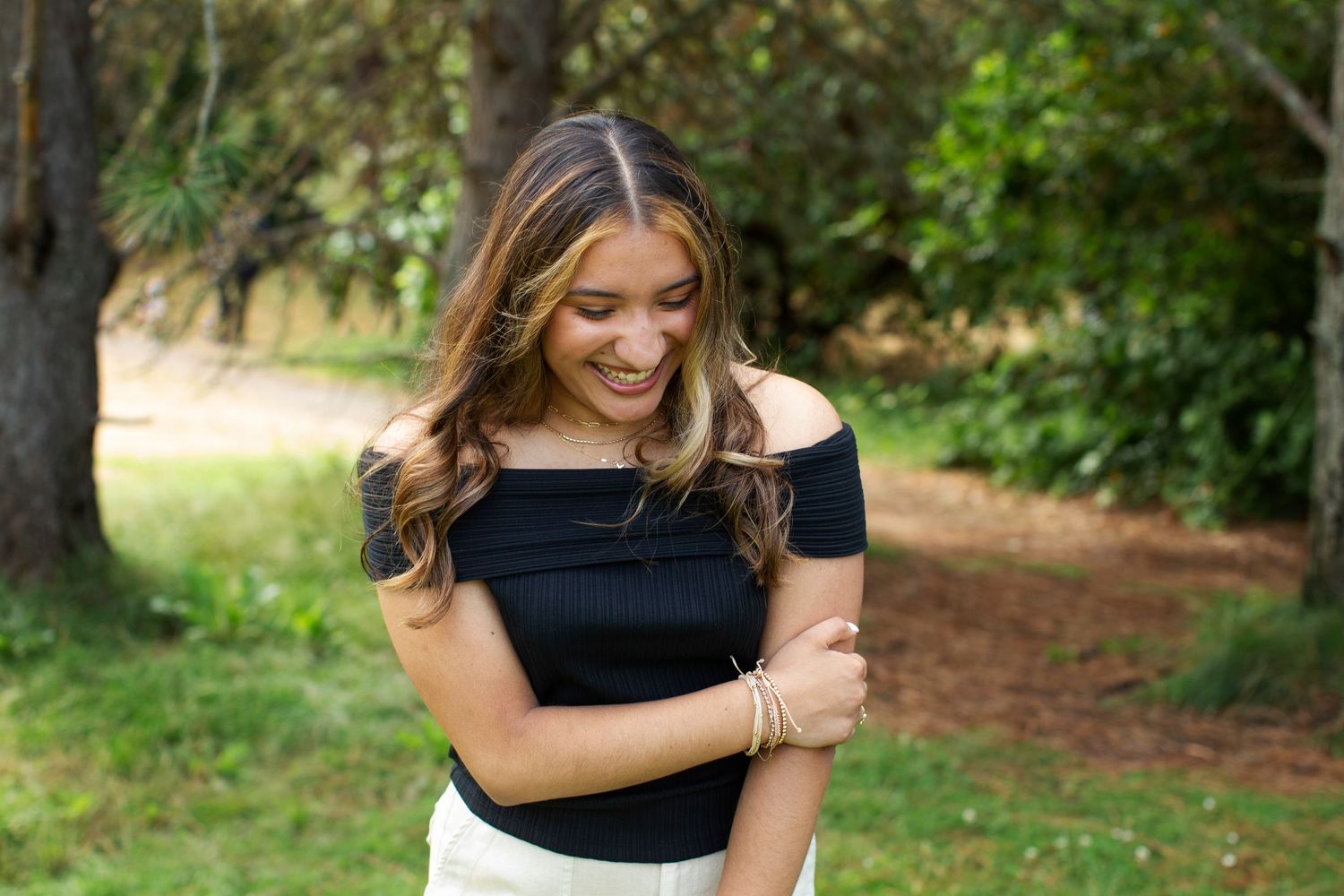 Young woman in black off-shoulder top smiling downward while standing on a lush green garden path.