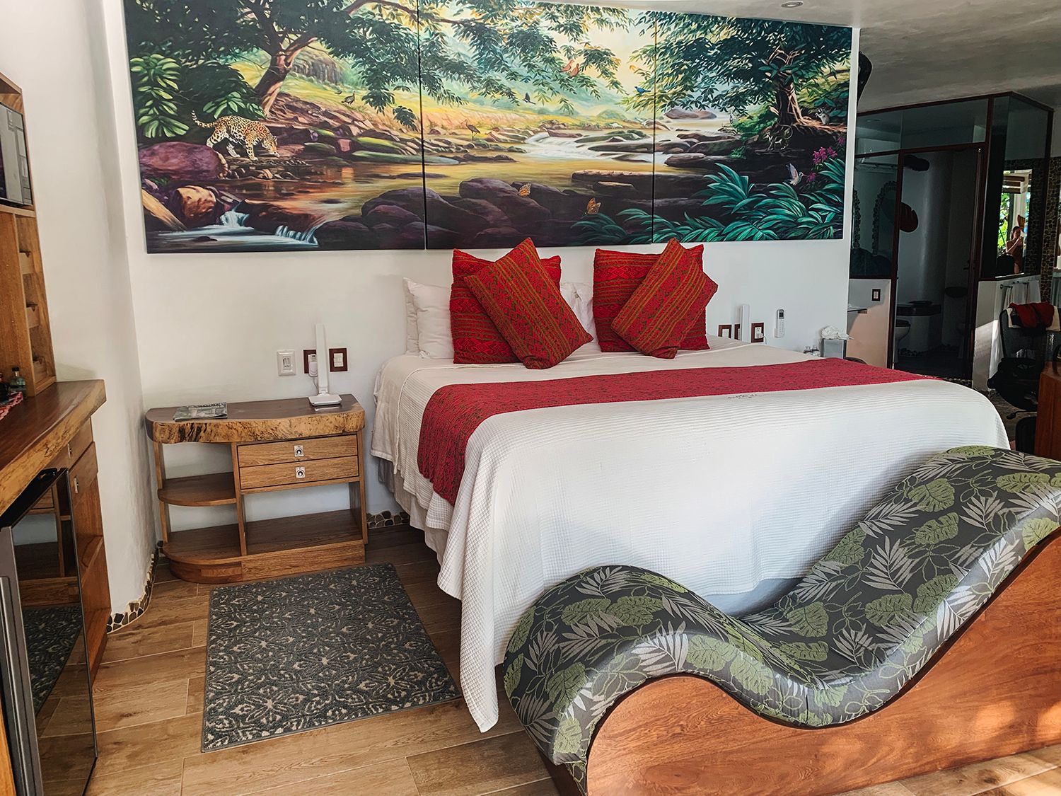 Cozy hotel room with vibrant jungle mural, red accent pillows, white bedding, and wooden furnishings.