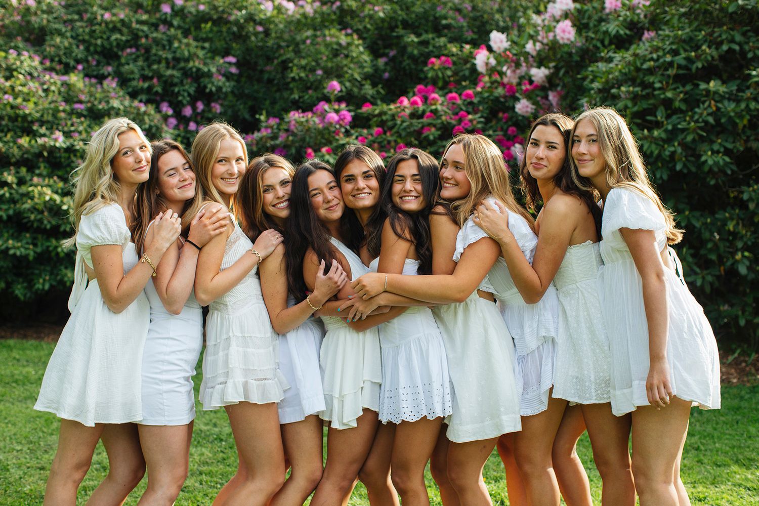 Group of young women in white dresses posing together outdoors with blooming pink flowers in the background.