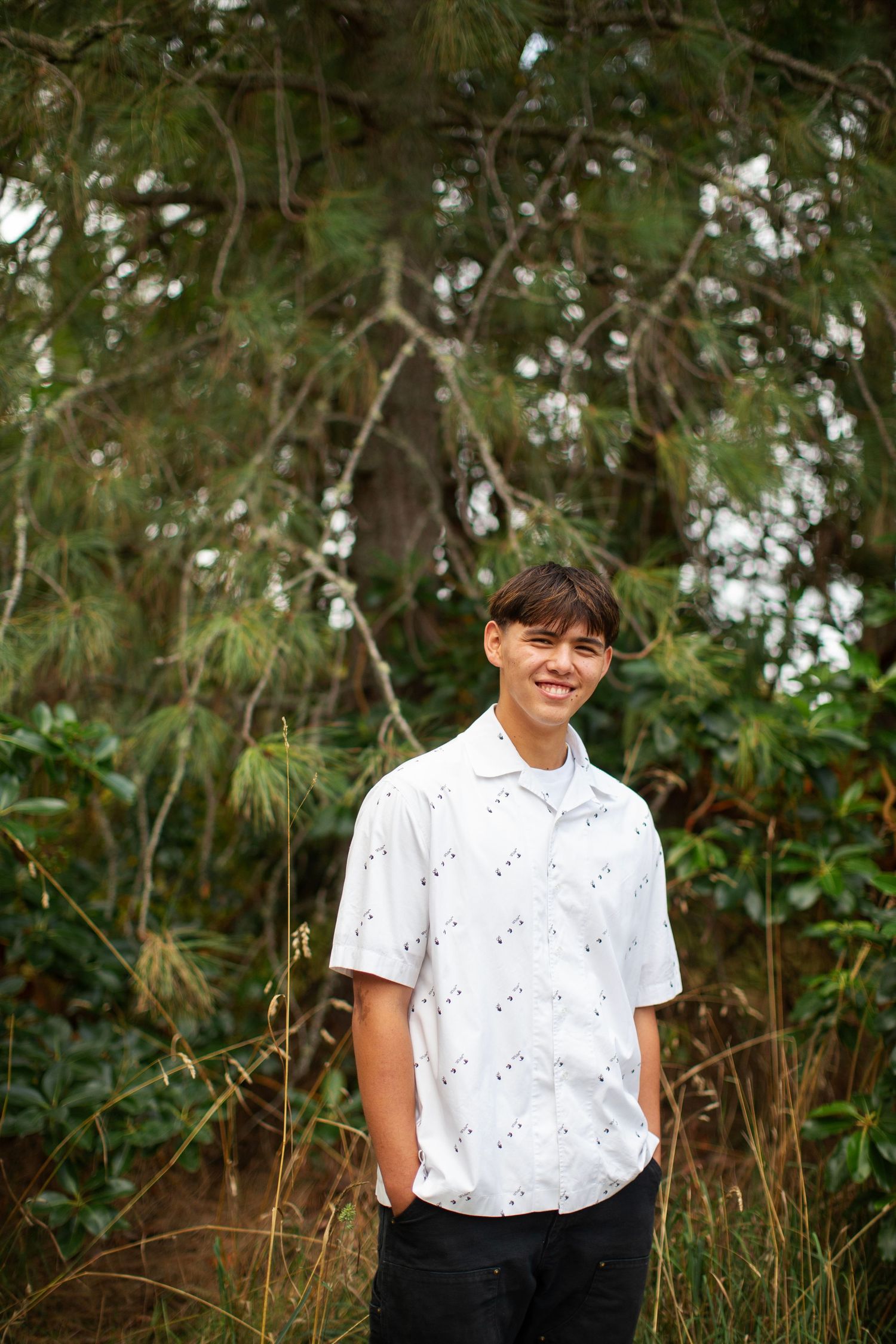 Smiling teen boy in white patterned shirt standing in front of lush green pine trees.