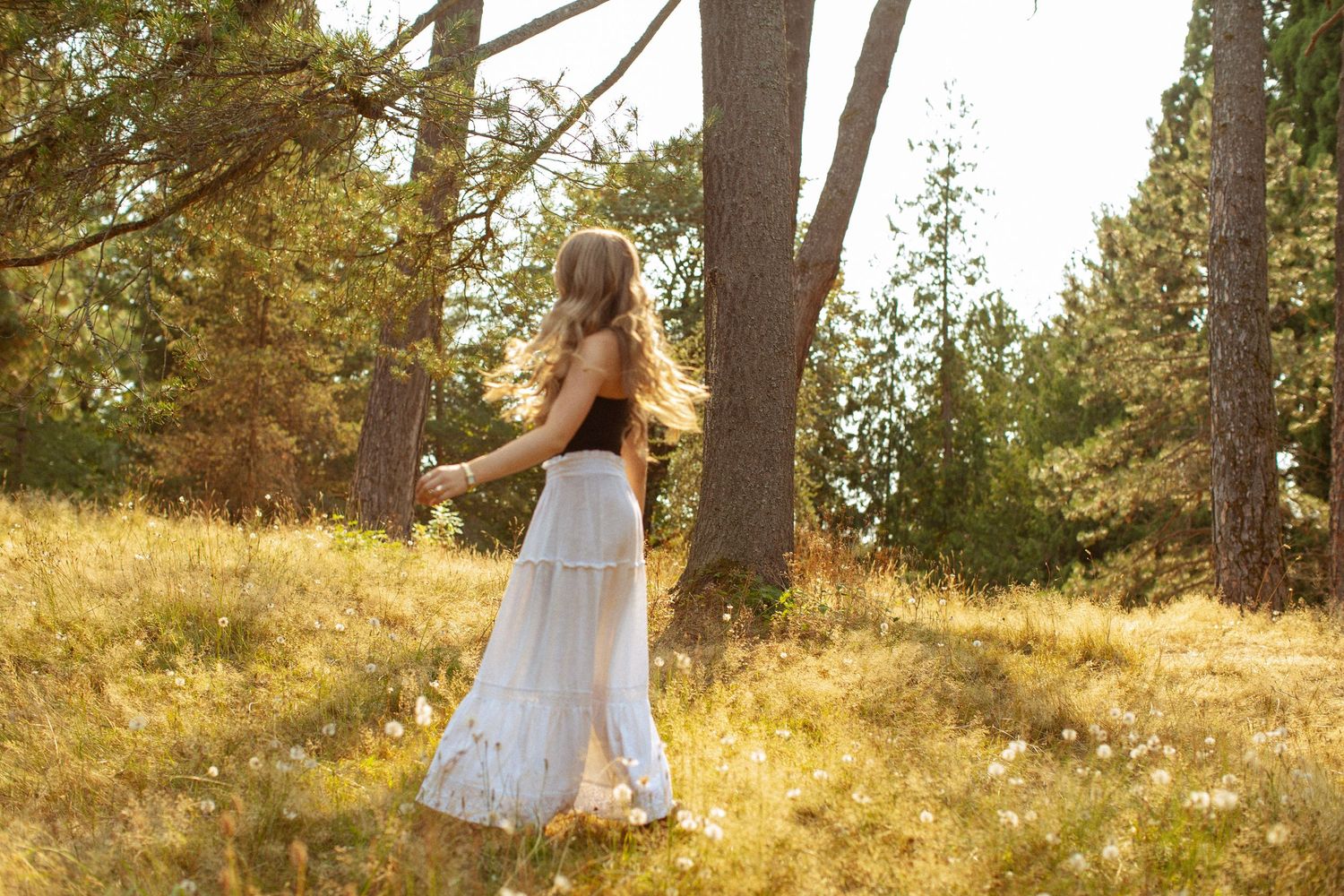 Young woman in white maxi skirt and black top walking through sunlit forest meadow with tall pine trees.