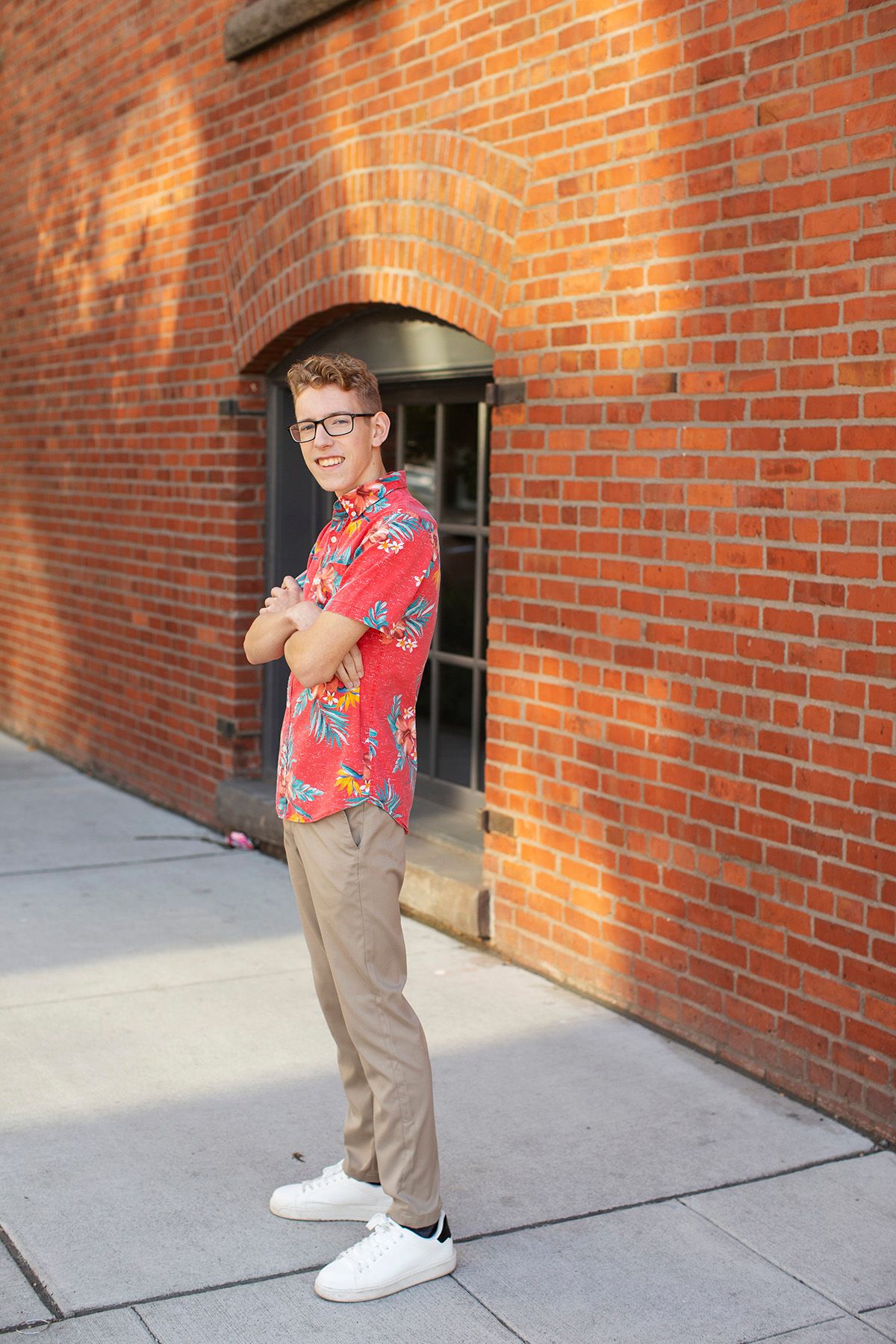 Young man in red floral shirt and khaki pants standing confidently against a red brick wall.